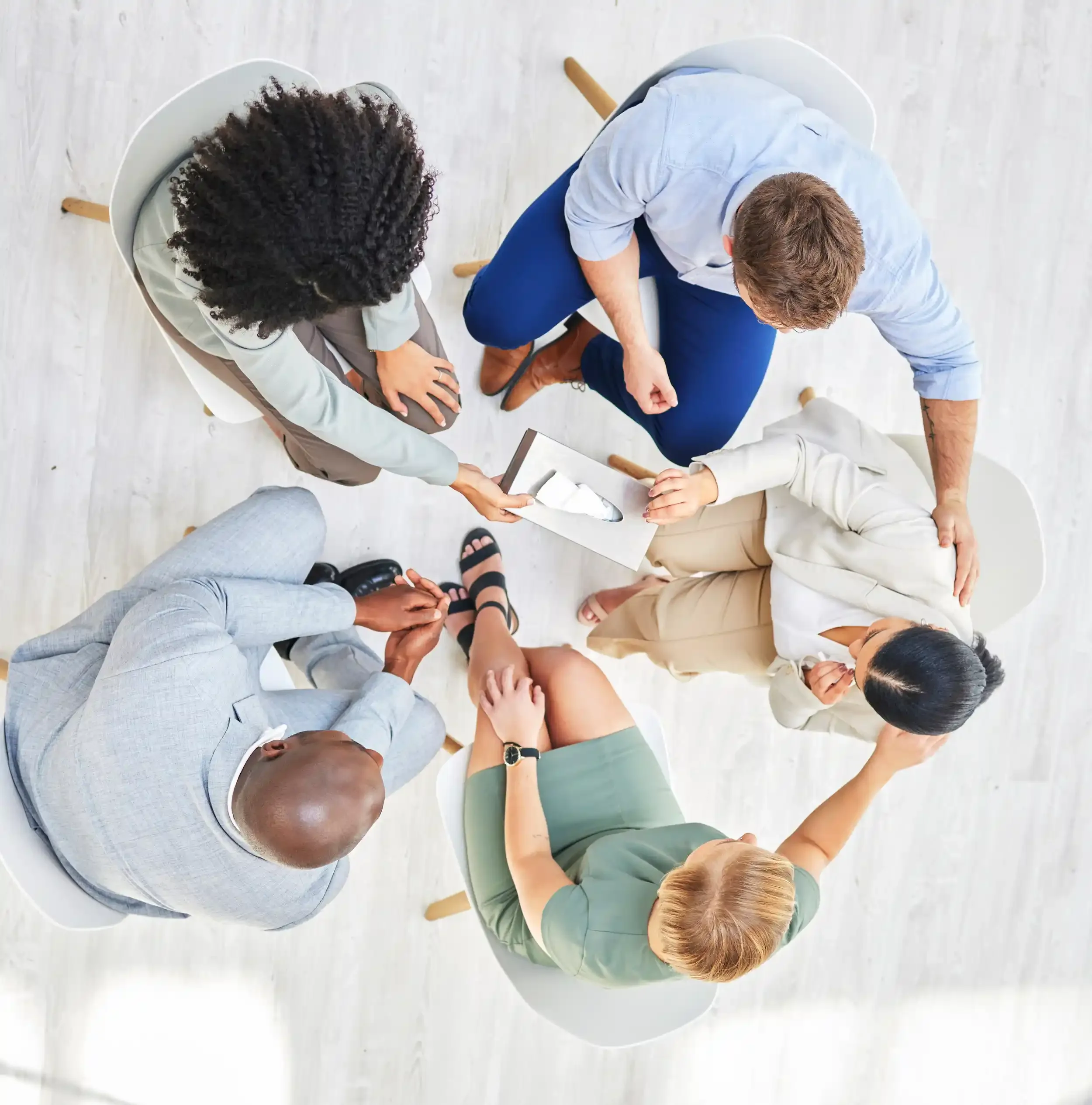 People sitting in a circle around a white table, engaged in a discussion, with some taking notes or holding papers, in a bright, minimalist room.