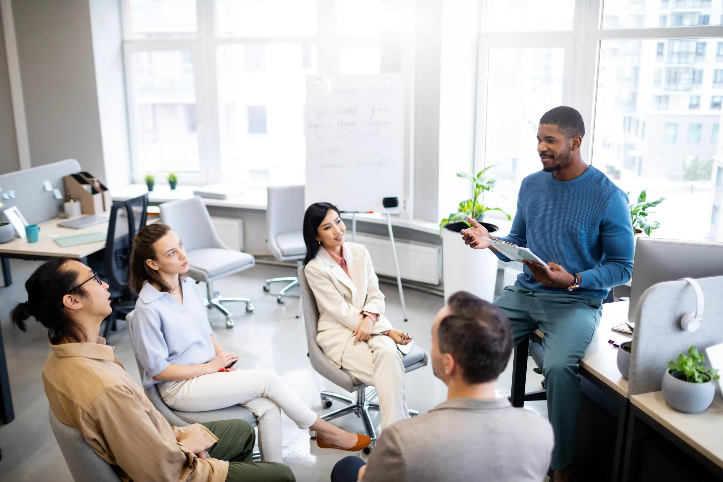 A group of people attending a presentation in a bright office space, with a man in a blue sweater standing and speaking while holding a clipboard.
