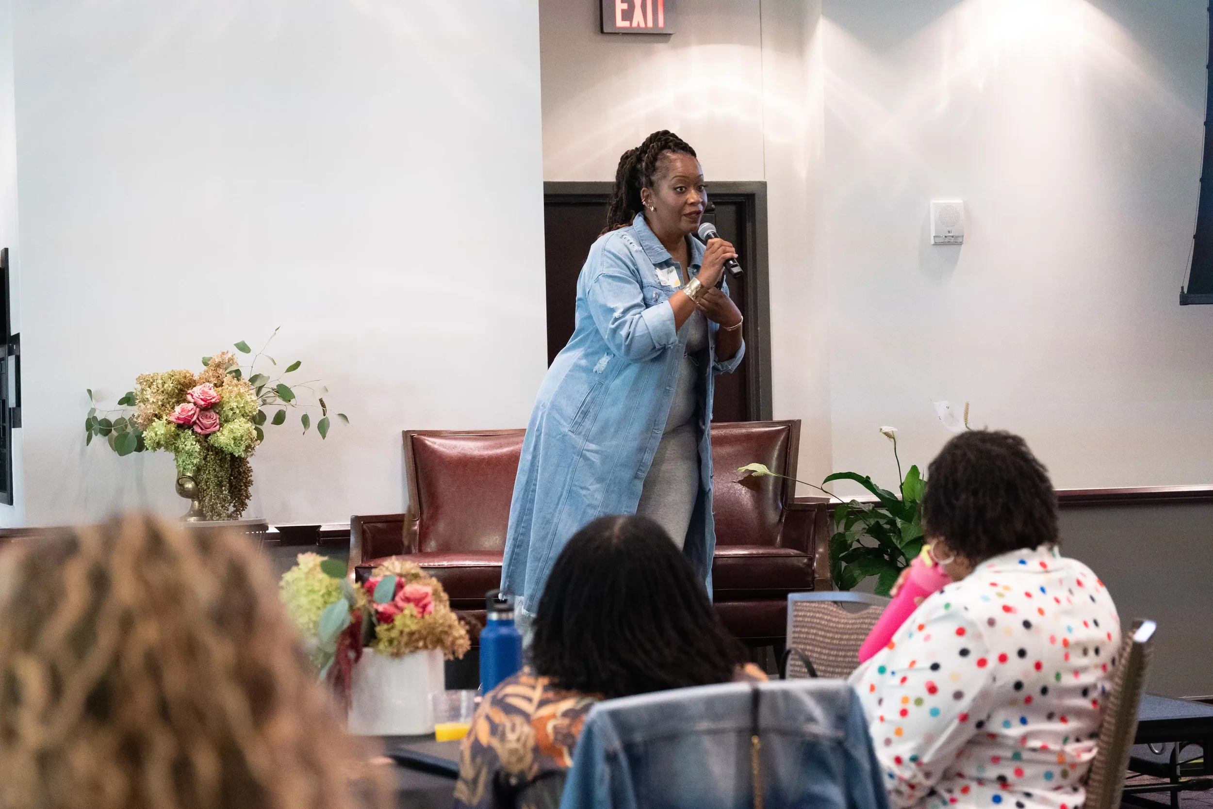 A woman in a denim jacket speaking into a microphone during a presentation or event, surrounded by seated children and colorful flower arrangements, in a room with beige walls and an 'EXIT' sign above the door.