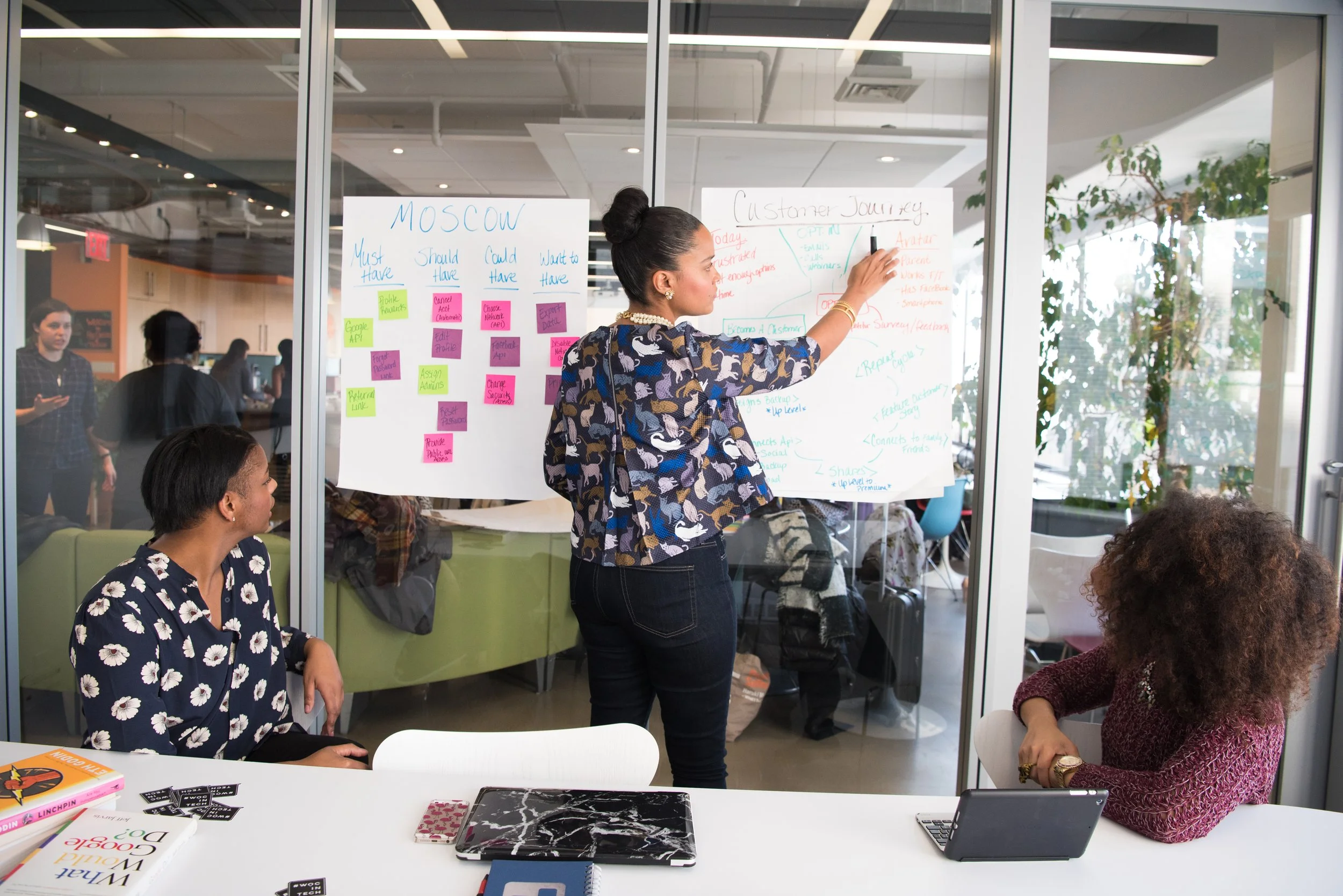 A woman in a patterned shirt is writing on a large whiteboard with notes and diagrams in a modern office or conference room, while others are seated and standing nearby, engaging in a discussion.