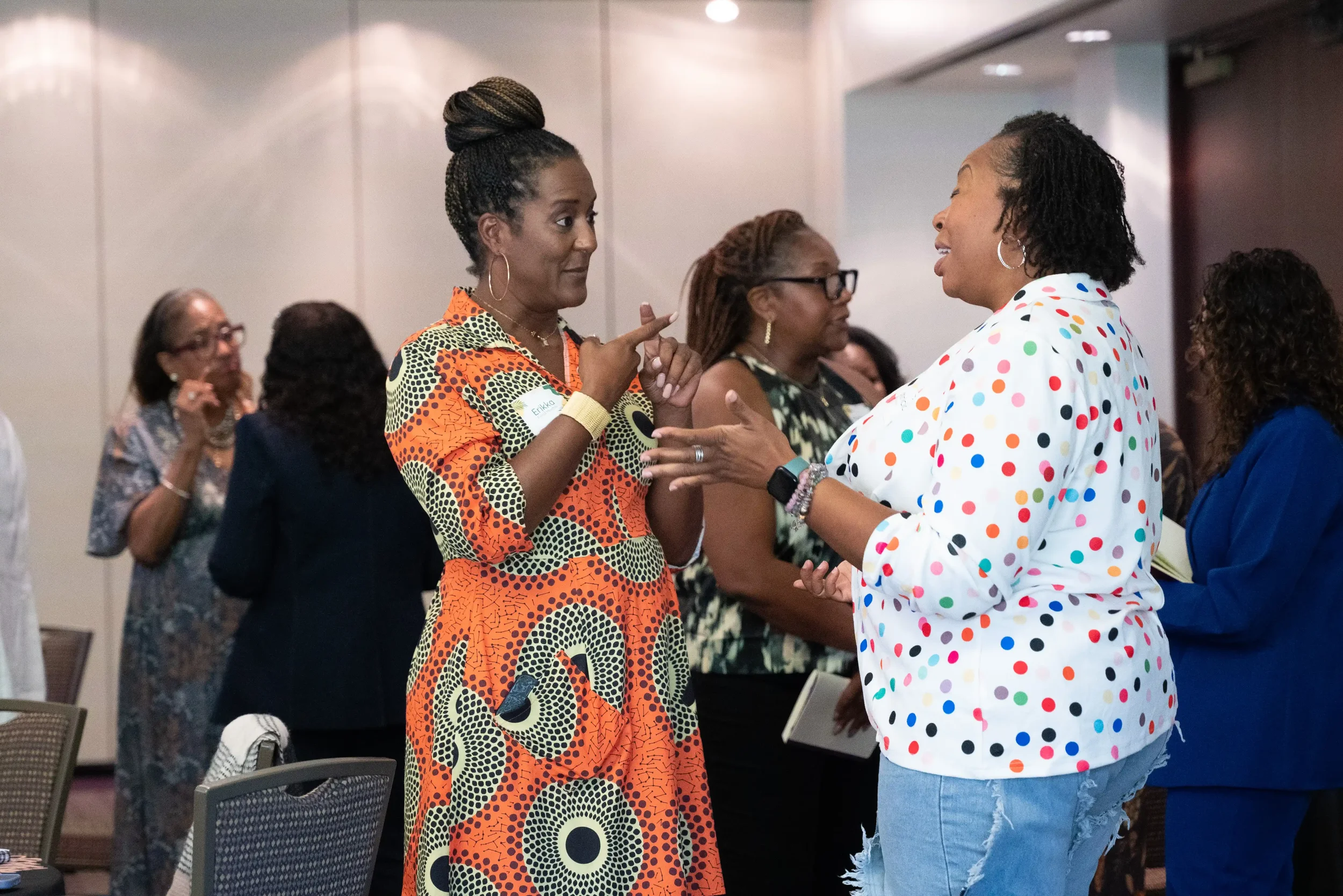 Two women are engaged in conversation at a social gathering, with one wearing an orange patterned dress and the other in a white shirt with colorful polka dots. Several other women are in the background, talking.