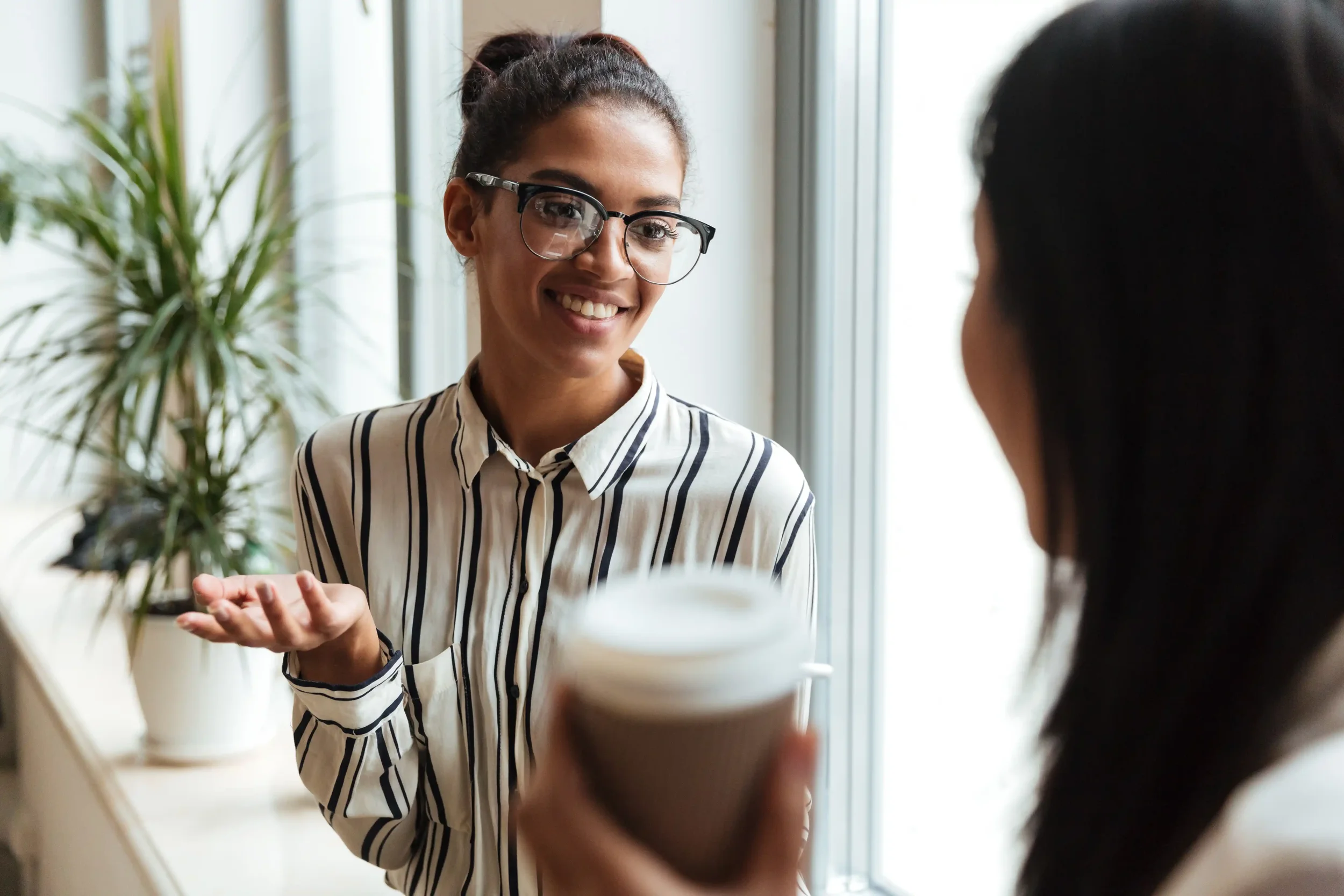 A woman with glasses smiling and talking to another person in an office setting, with a potted plant nearby.