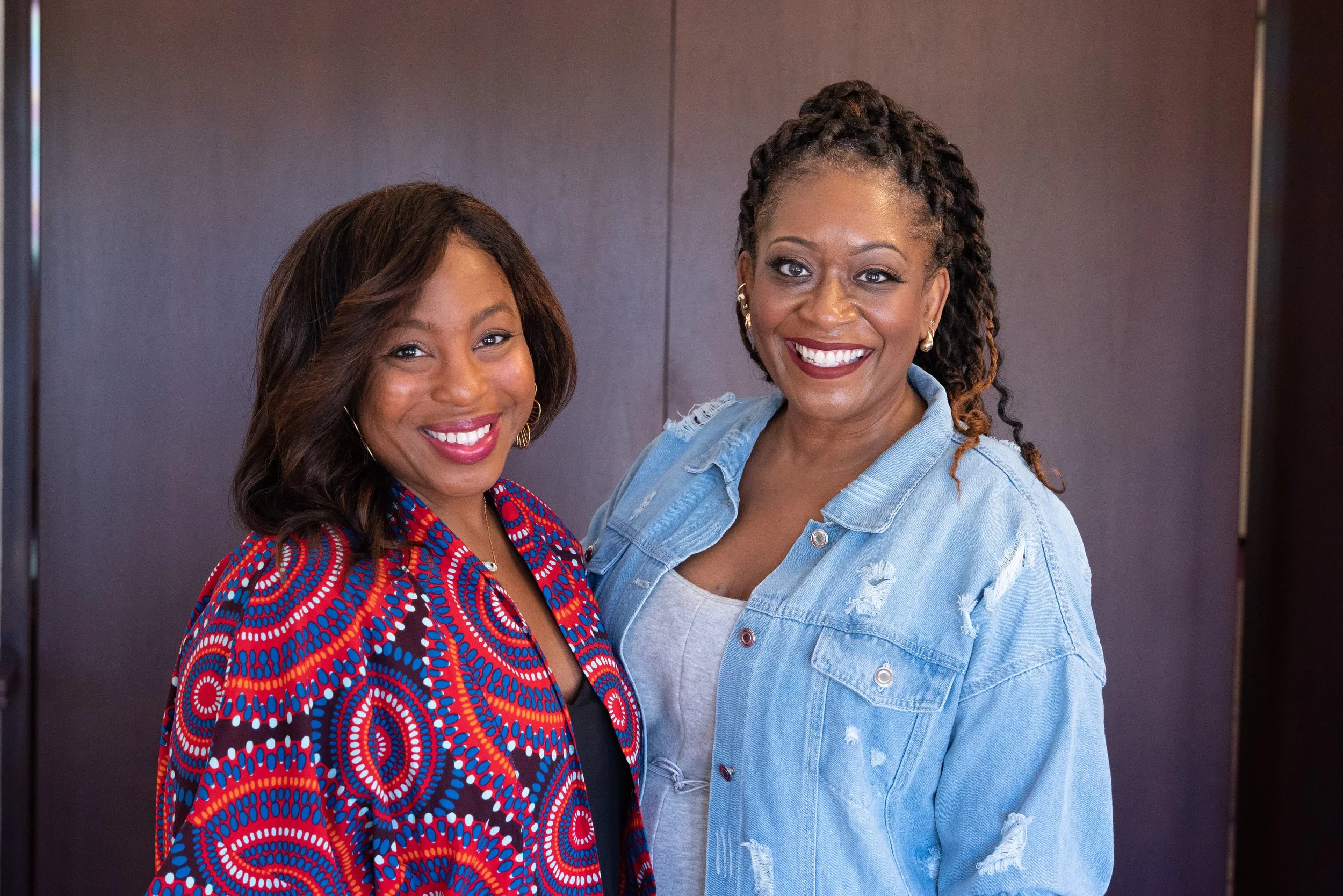 Two smiling women standing close together indoors against a plain dark background, one wearing a vibrant red, blue, and black patterned dress, and the other wearing a light blue denim jacket over a white top with distressed details.