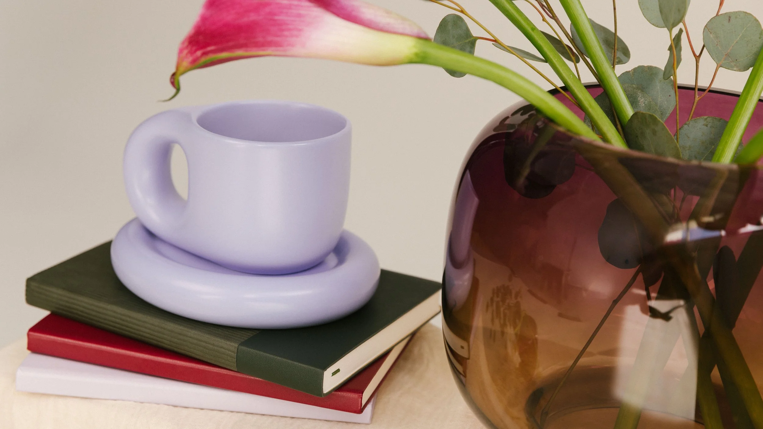 A still life image showing a white ceramic coffee mug resting on top of a purple ceramic saucer, placed on top of two closed books with green and red covers. To the right, there is a large, semi-transparent, brown glass vase containing green leafy stems and a partially visible pink flower.