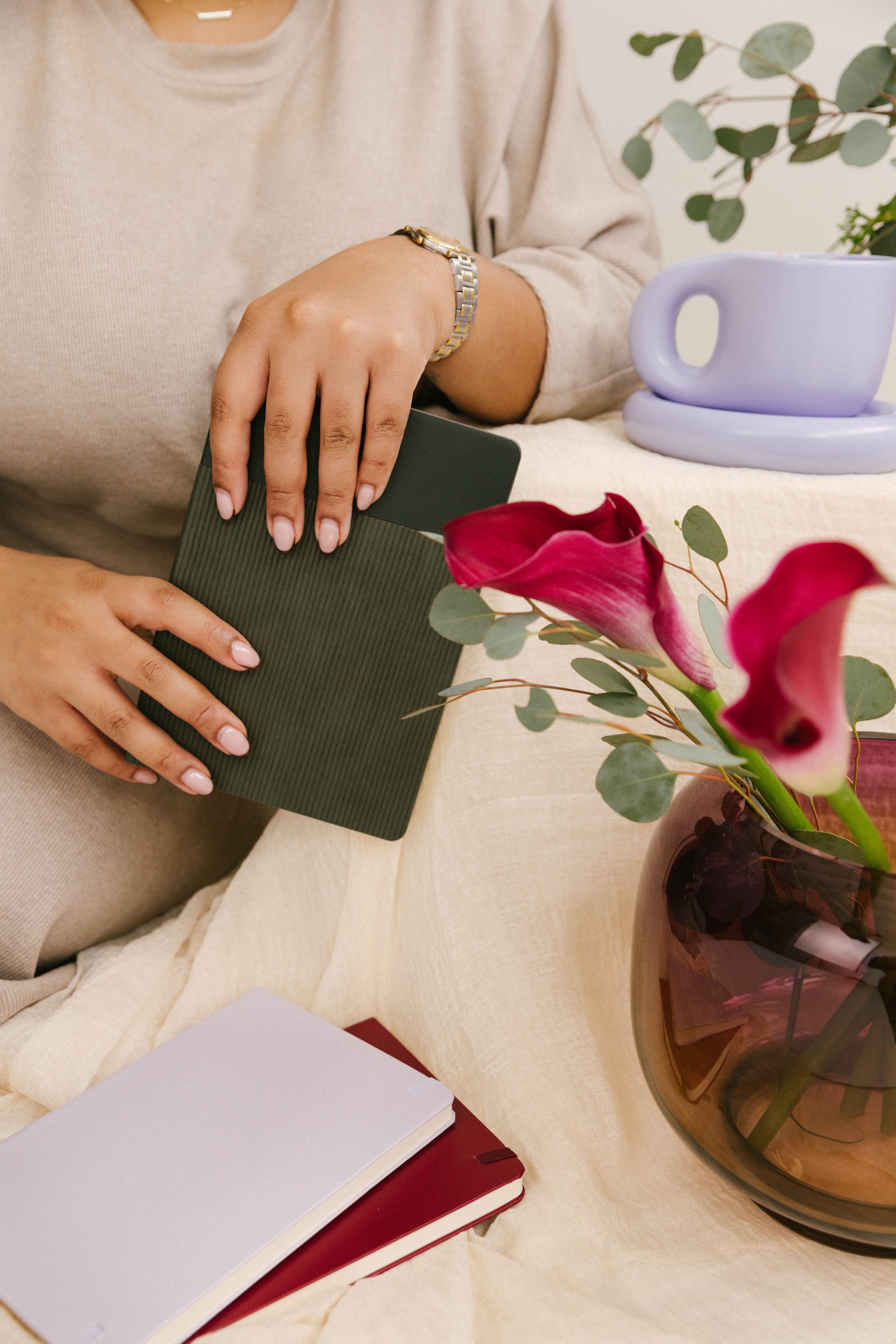 Person placing a book into a dark green shelf on a table with a purple mug, a vase of pink calla lilies, two notebooks, and a plant in the background.
