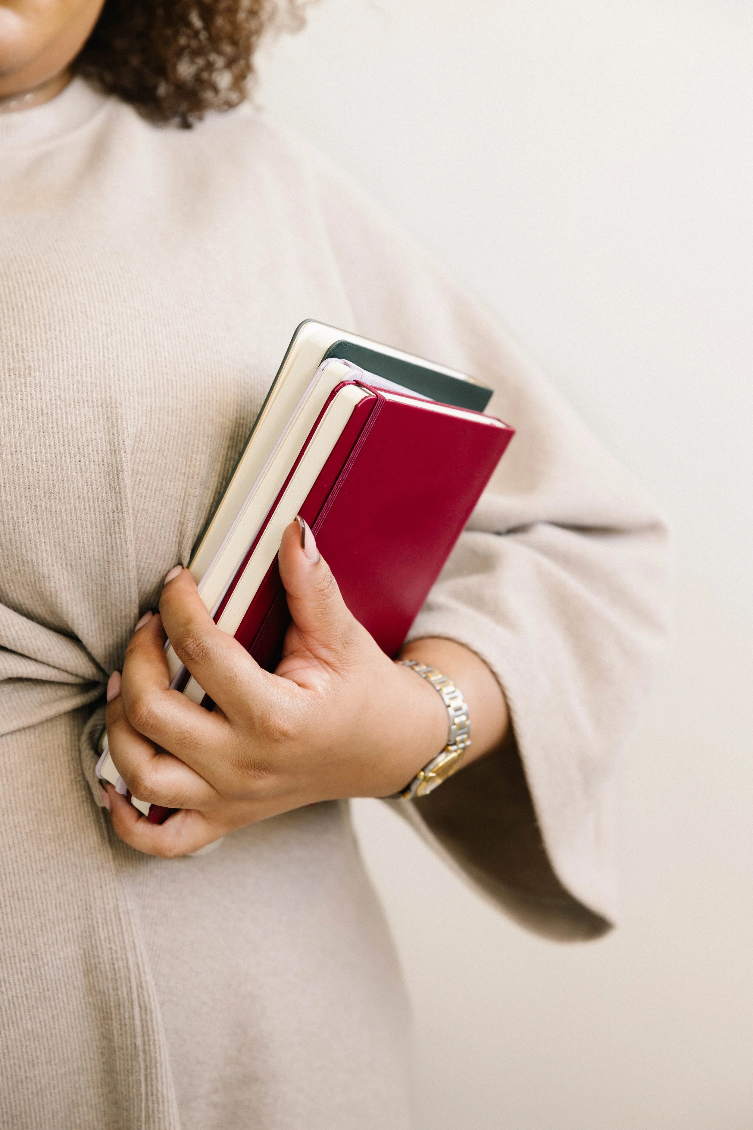 A woman holding a stack of notebooks or planners, standing against a plain background, wearing a beige sweater and a gold watch.