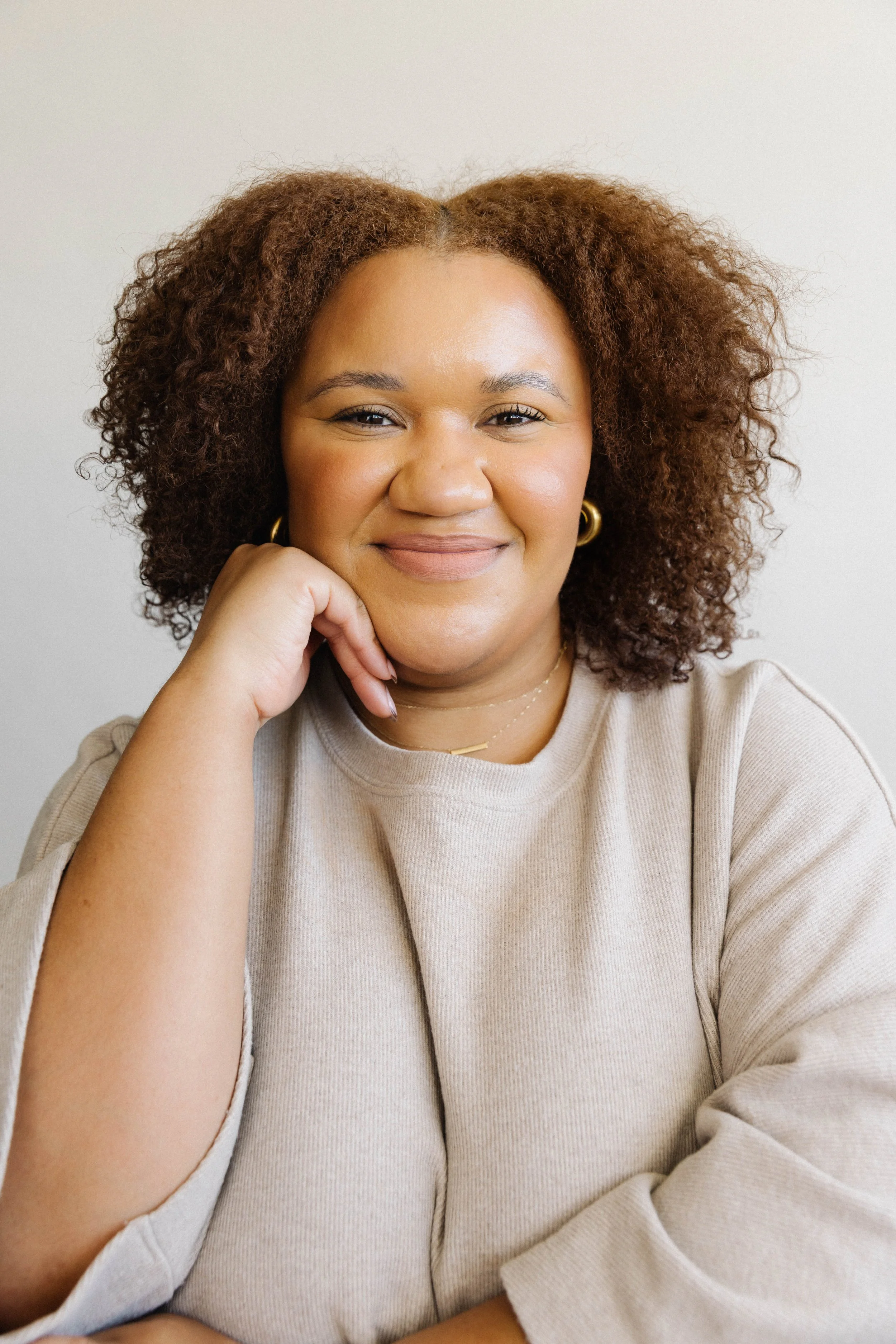A woman with curly brown hair smiling and resting her chin on her hand, wearing a beige top and gold jewelry.