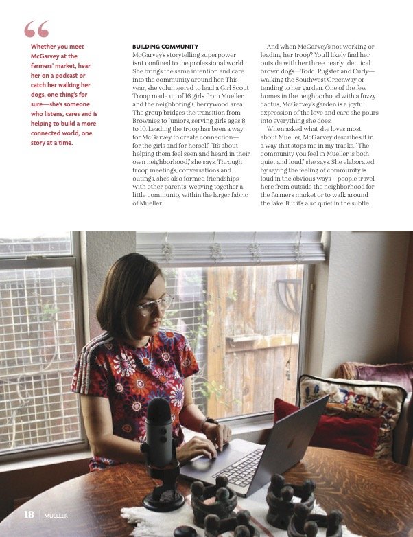 Woman with glasses, wearing a red floral shirt, sitting at wooden table with laptop and microphone, by window with wooden blinds. Small figurines on table.