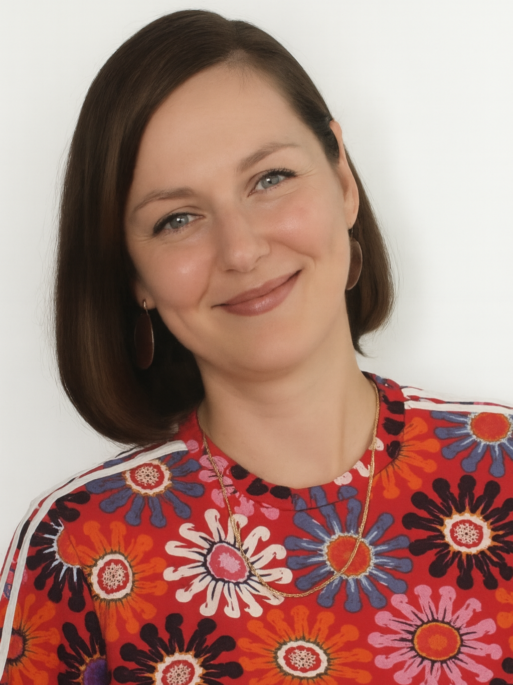 A woman with brown hair, blue eyes, and a warm smile, wearing a colorful floral top and jewelry, standing against a plain background.