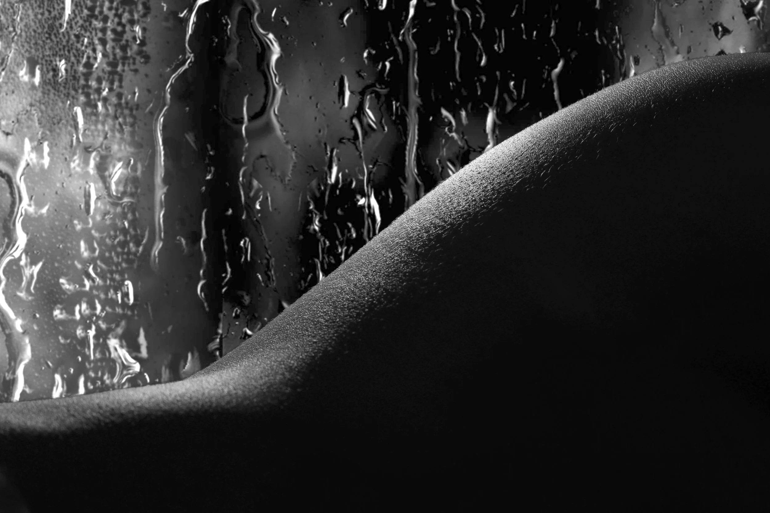 Close-up of raindrops on a window with a smooth skin in the foreground, in black and white.