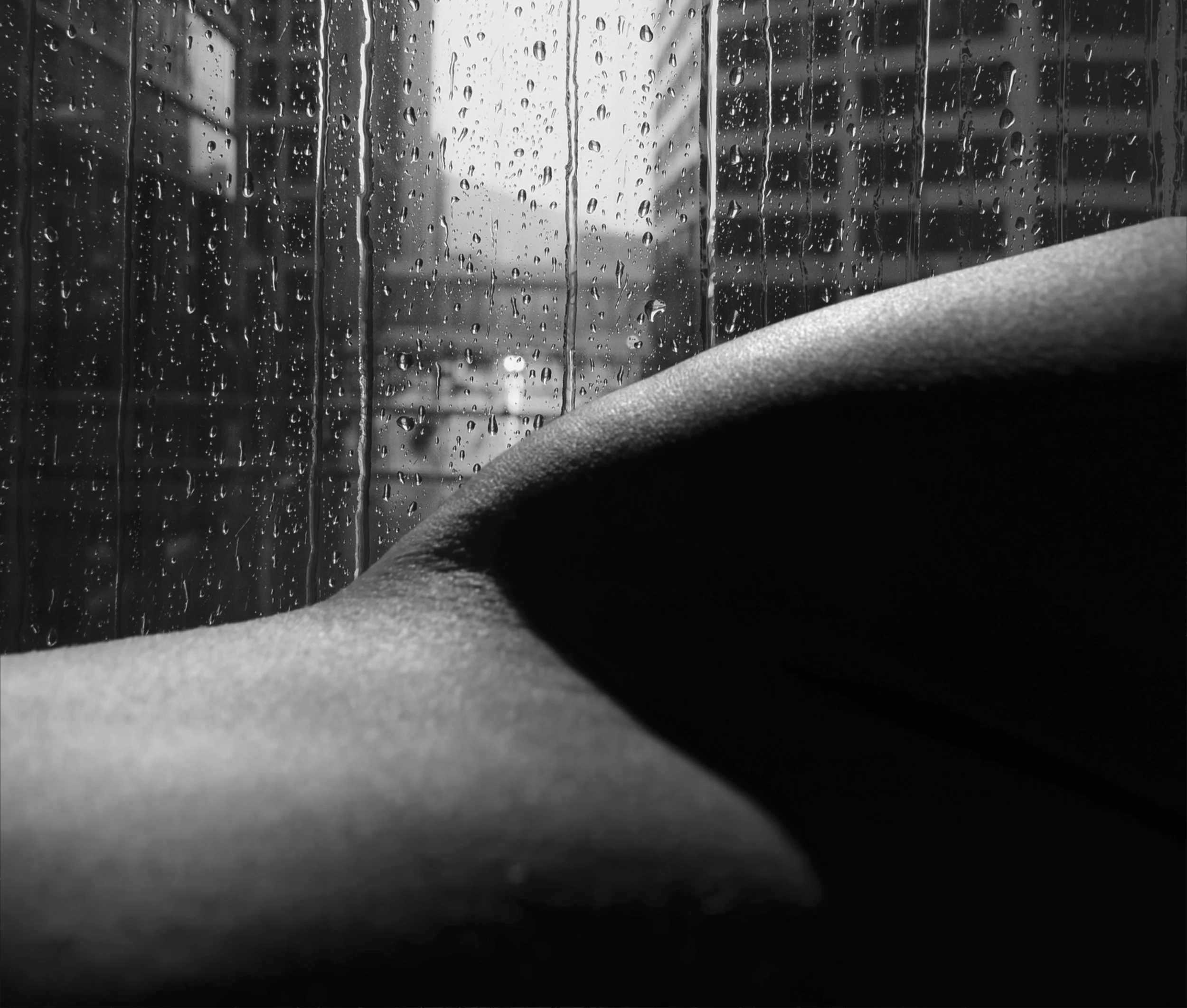 A black and white photo taken of body in front of raindrops on a window and a city scape.
