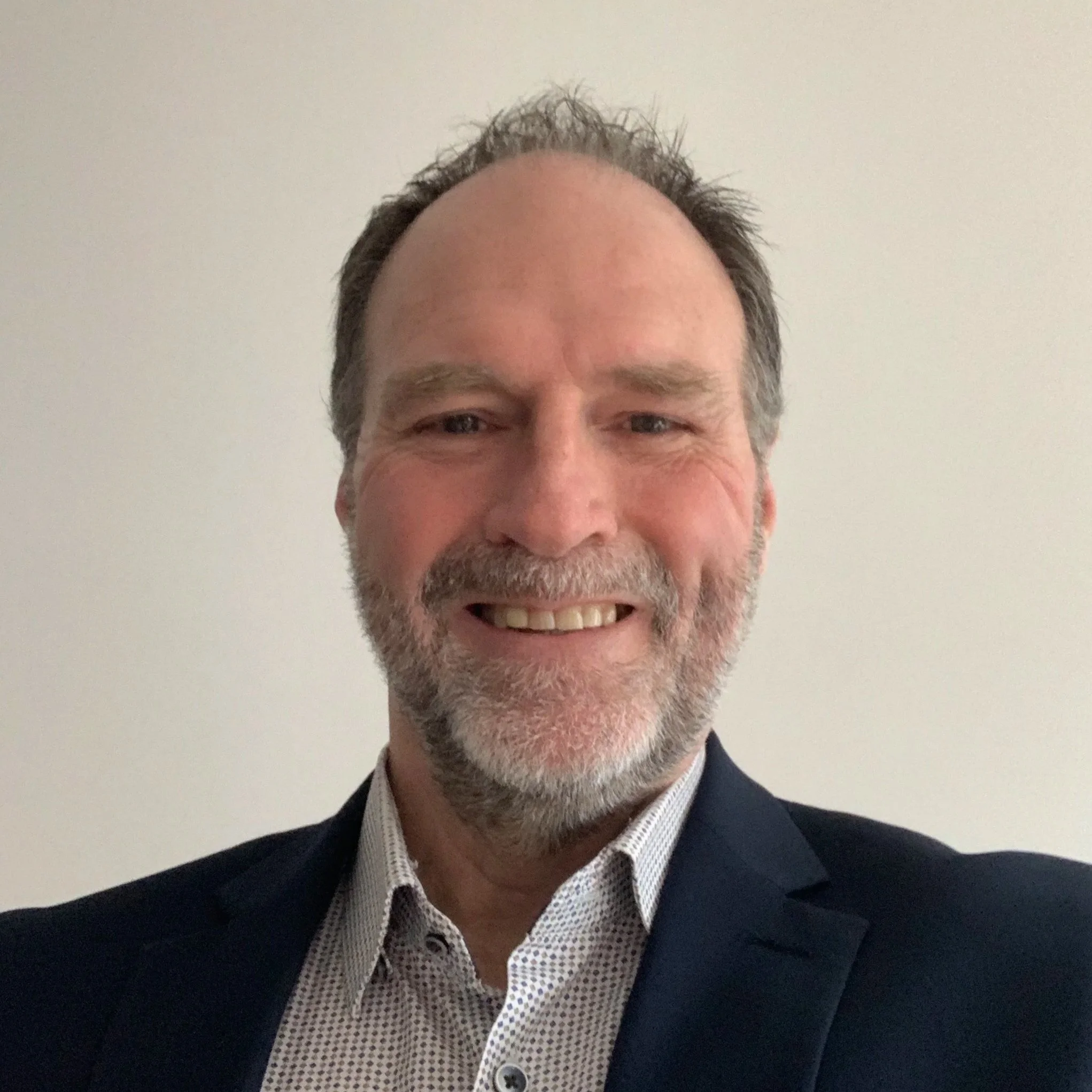 A smiling middle-aged man with gray hair and a beard, wearing a dark blazer and a collared shirt, standing against a plain white wall.