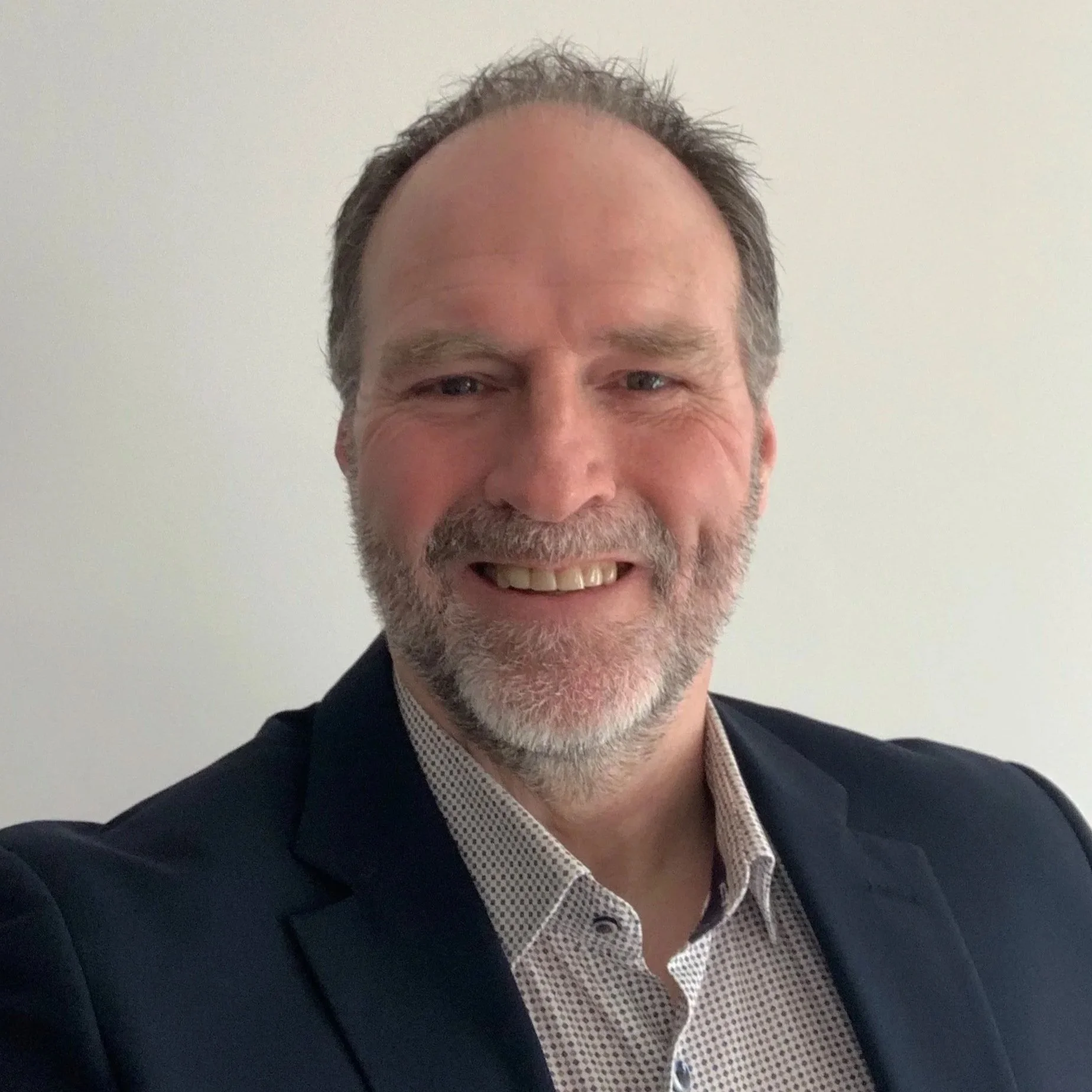 A middle-aged man with a beard and short hair smiling and wearing a dark blazer over a light patterned shirt.