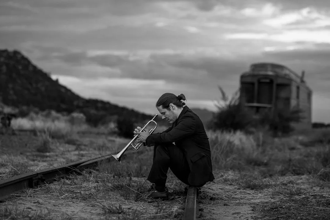 A man in a black suit sitting on railroad tracks, playing a trumpet, with an abandoned train in the background, black and white photo.