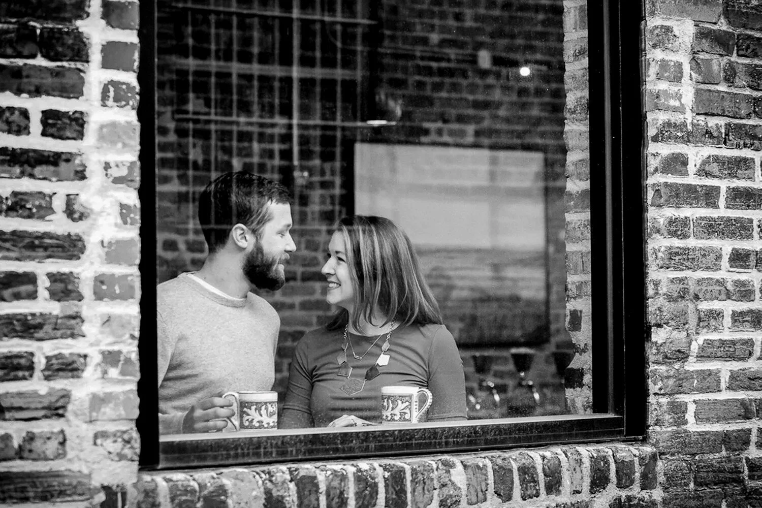 A man and woman smiling at each other through a cafe window, holding mugs, inside a brick-walled setting.