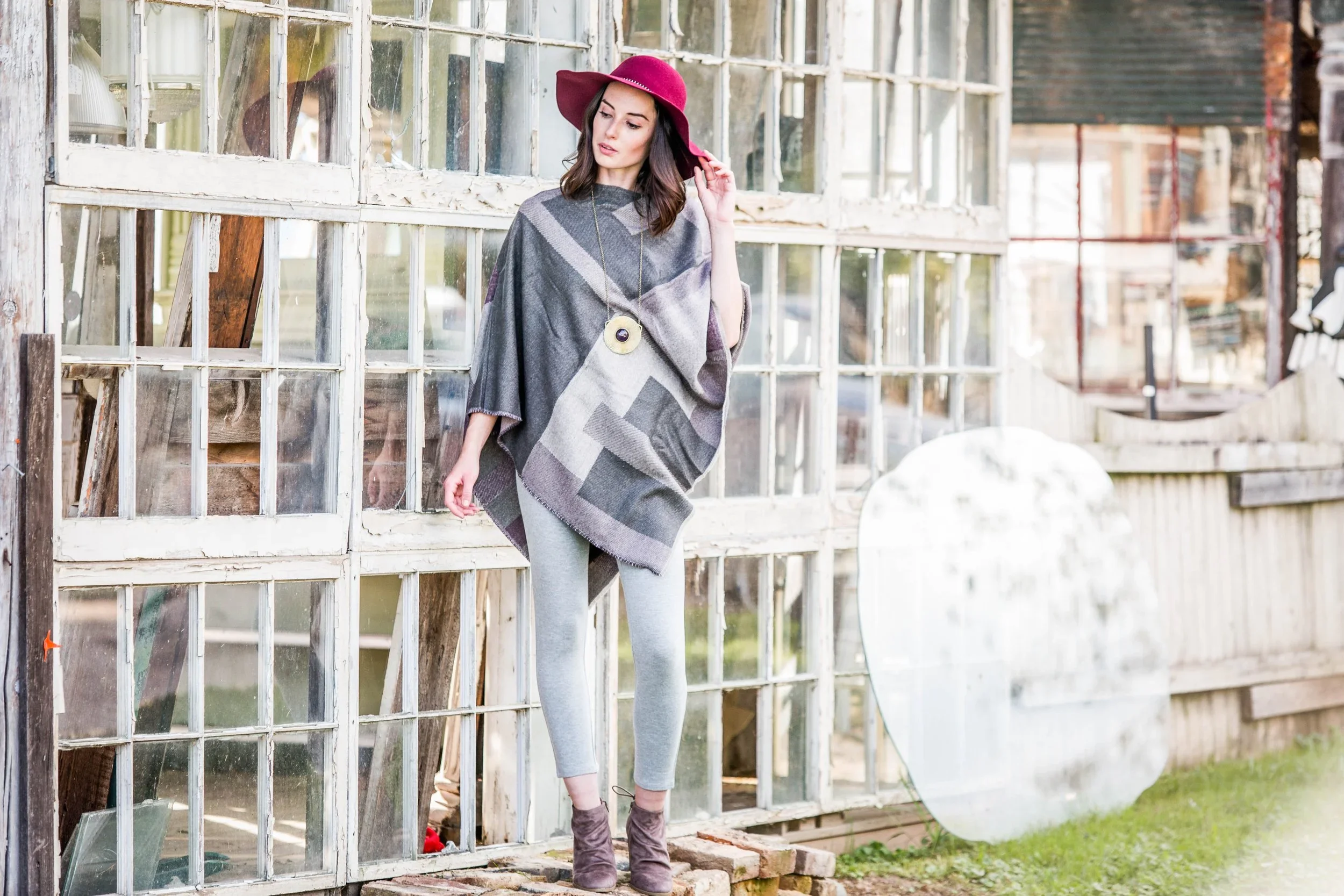 A woman wearing a gray poncho, gray leggings, and ankle boots stands outside next to a broken glass greenhouse wall, holding a pink wide-brim hat and looking down 