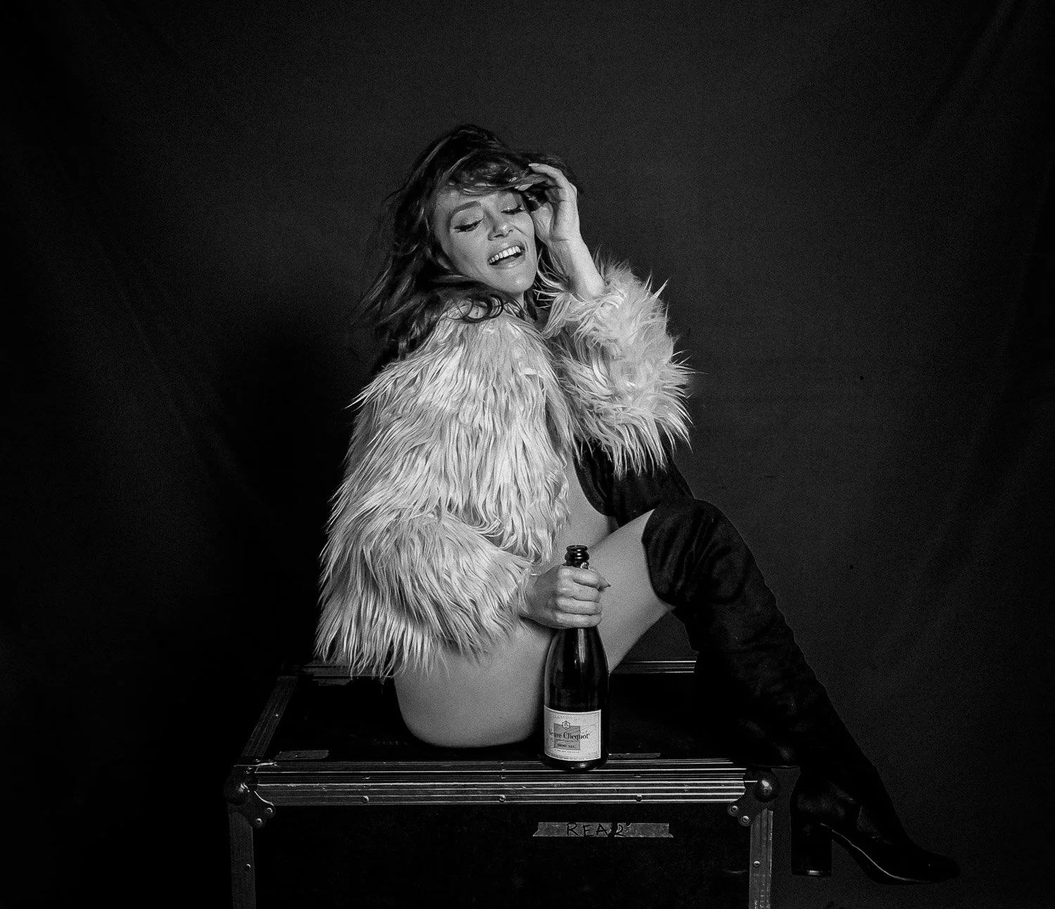 Editorial Black and white photo of a woman with wavy hair, wearing a fluffy jacket, sitting on a small table with one leg up, holding a bottle of wine, against a dark background.