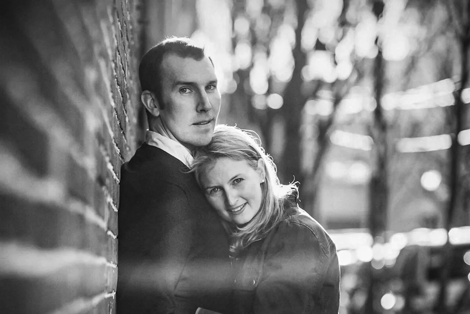 A black and white photo of a young man and woman leaning against a brick wall outdoors, with trees and blurred background behind them. The woman is smiling and cuddling close to the man, who is looking directly at the camera.