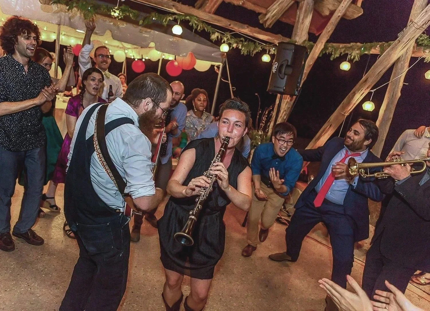 People dancing and playing instruments at a lively celebration or party, with decorations and string lights overhead.