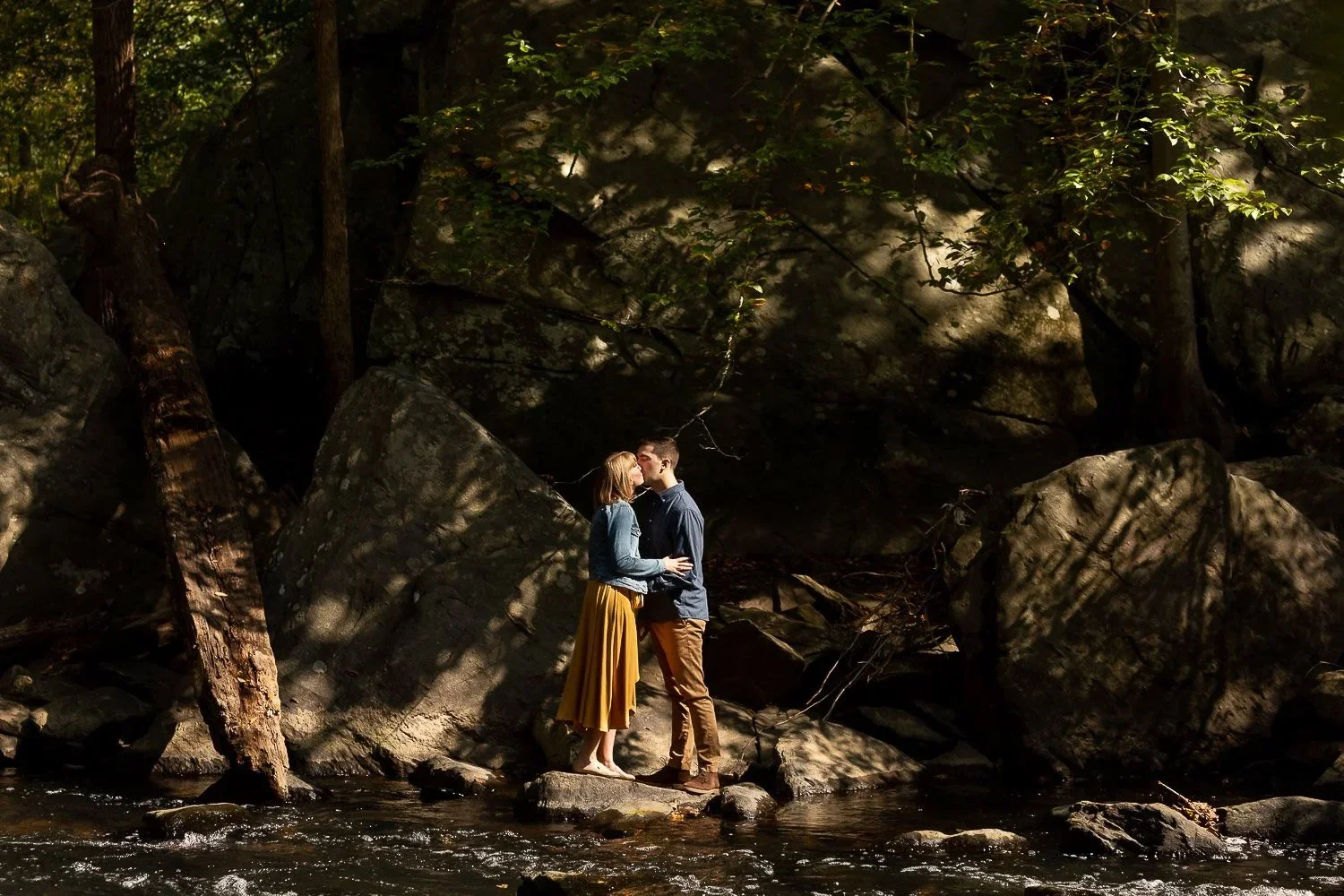 A couple stands on a rock in a stream, surrounded by large rocks and trees in a forest, as they share a kiss.