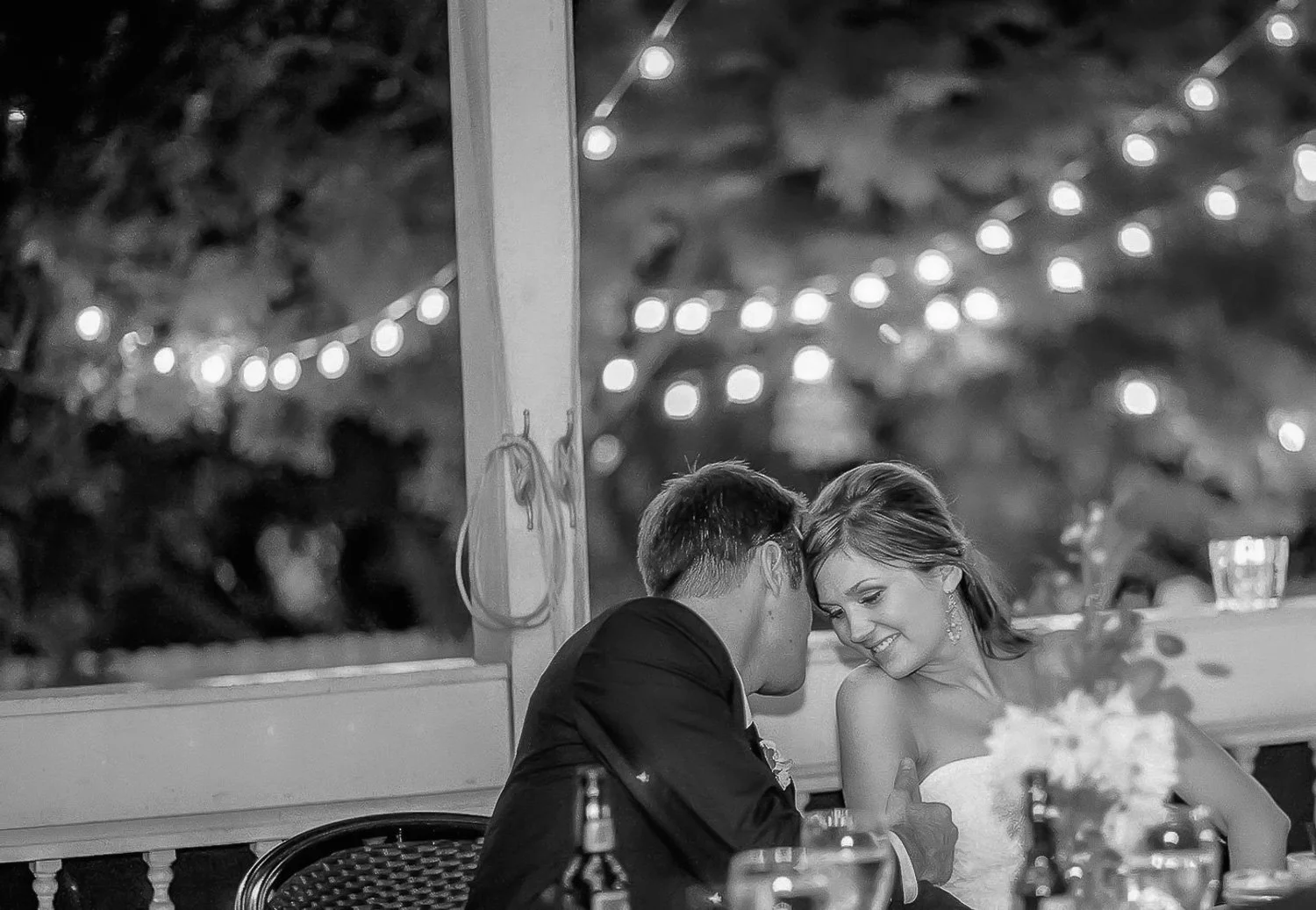A black and white photo of a couple at a wedding reception, sitting together at a table, leaning their foreheads and smiling at each other, with string lights in the background.
