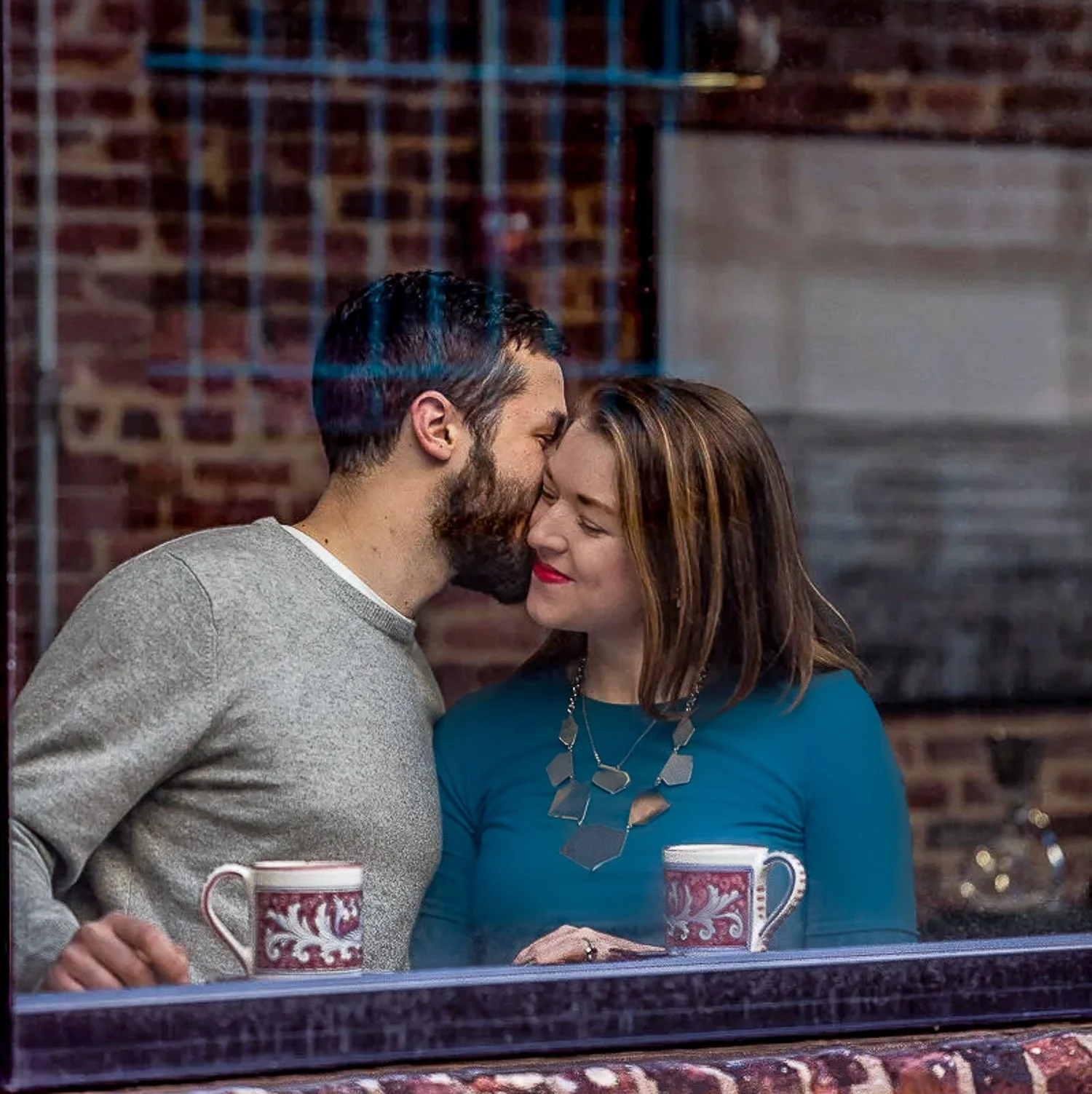 A couple sharing a tender moment, touching foreheads and smiling softly, standing in front of a brick wall, with two coffee mugs on a table in front of them.