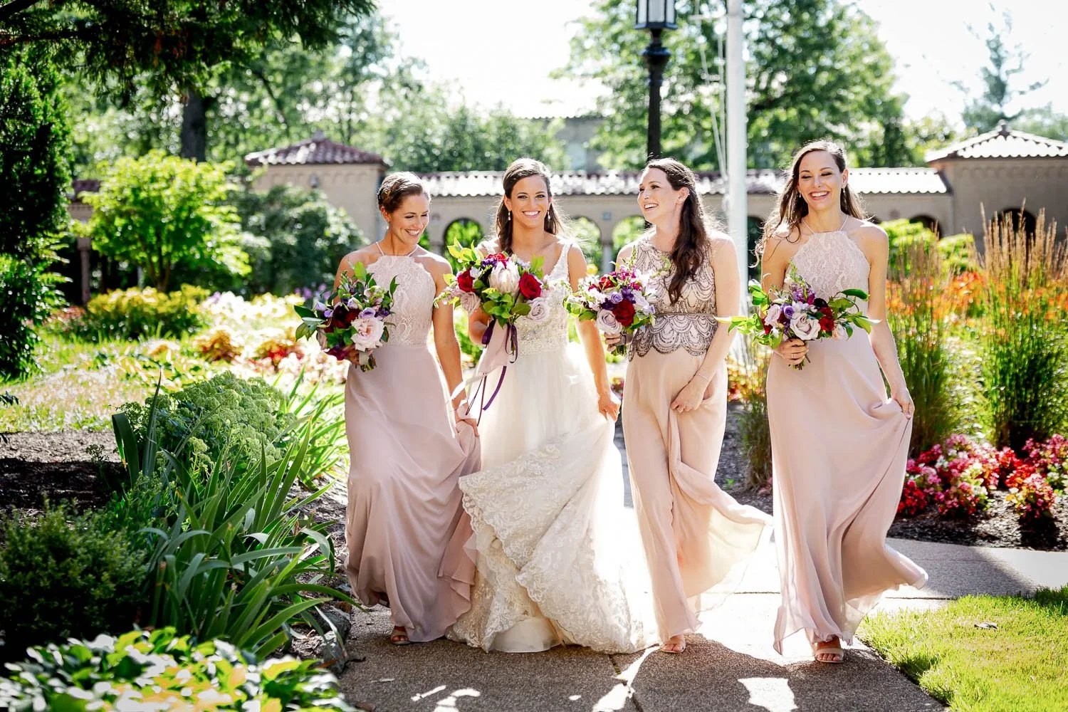 A bride and four bridesmaids walking through a garden on a sunny day, holding bouquets of flowers.