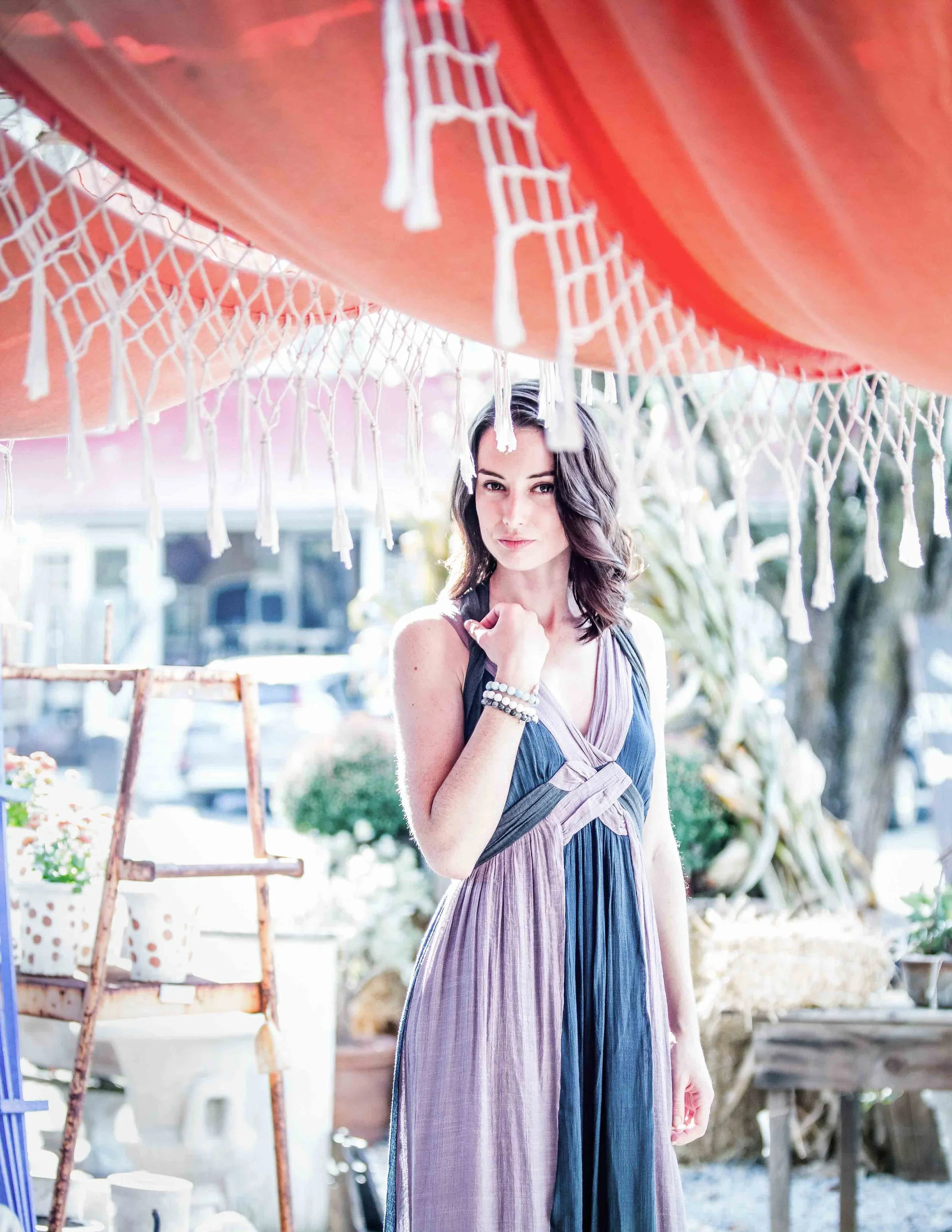 A woman in a sleeveless dress with a purple and blue color gradient standing outdoors under a red-orange umbrella with white tassels, with a background of greenery and outdoor furniture.
