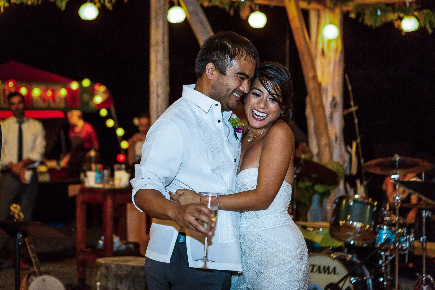 A smiling couple dancing at their wedding celebration, with the bride in a white lace dress holding a glass of champagne, and the groom in a white shirt, in an outdoor nighttime setting with string lights and a band in the background.