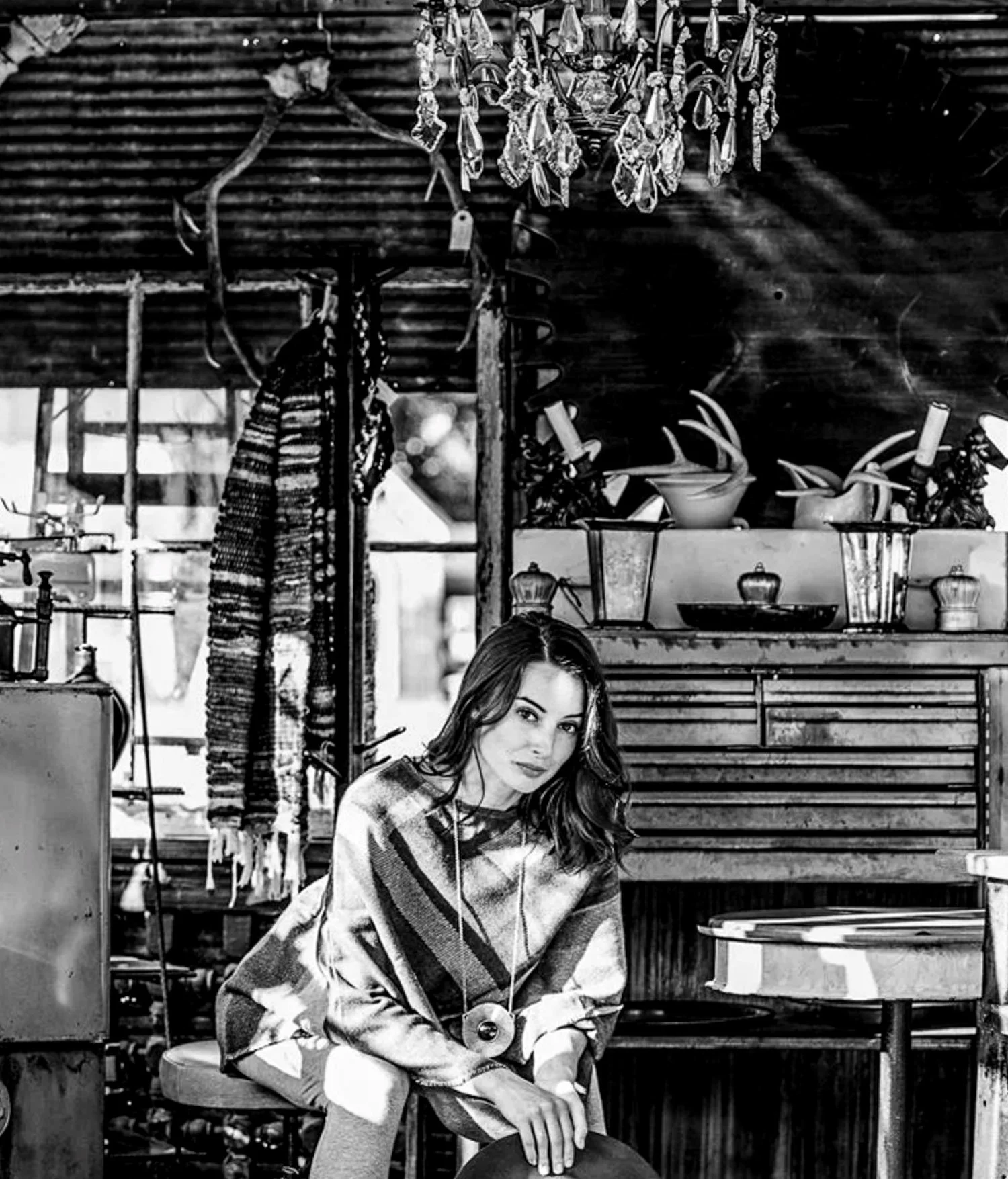 A young woman with long hair, wearing a plaid blanket, sits in a rustic wooden setting with antique items and decorative objects around her.