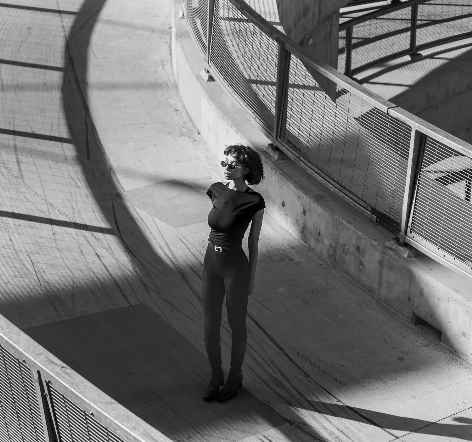 A woman wearing sunglasses, a black top, and black pants stands alone on an outdoor concrete walkway with shadows and a metal railing, in black and white.