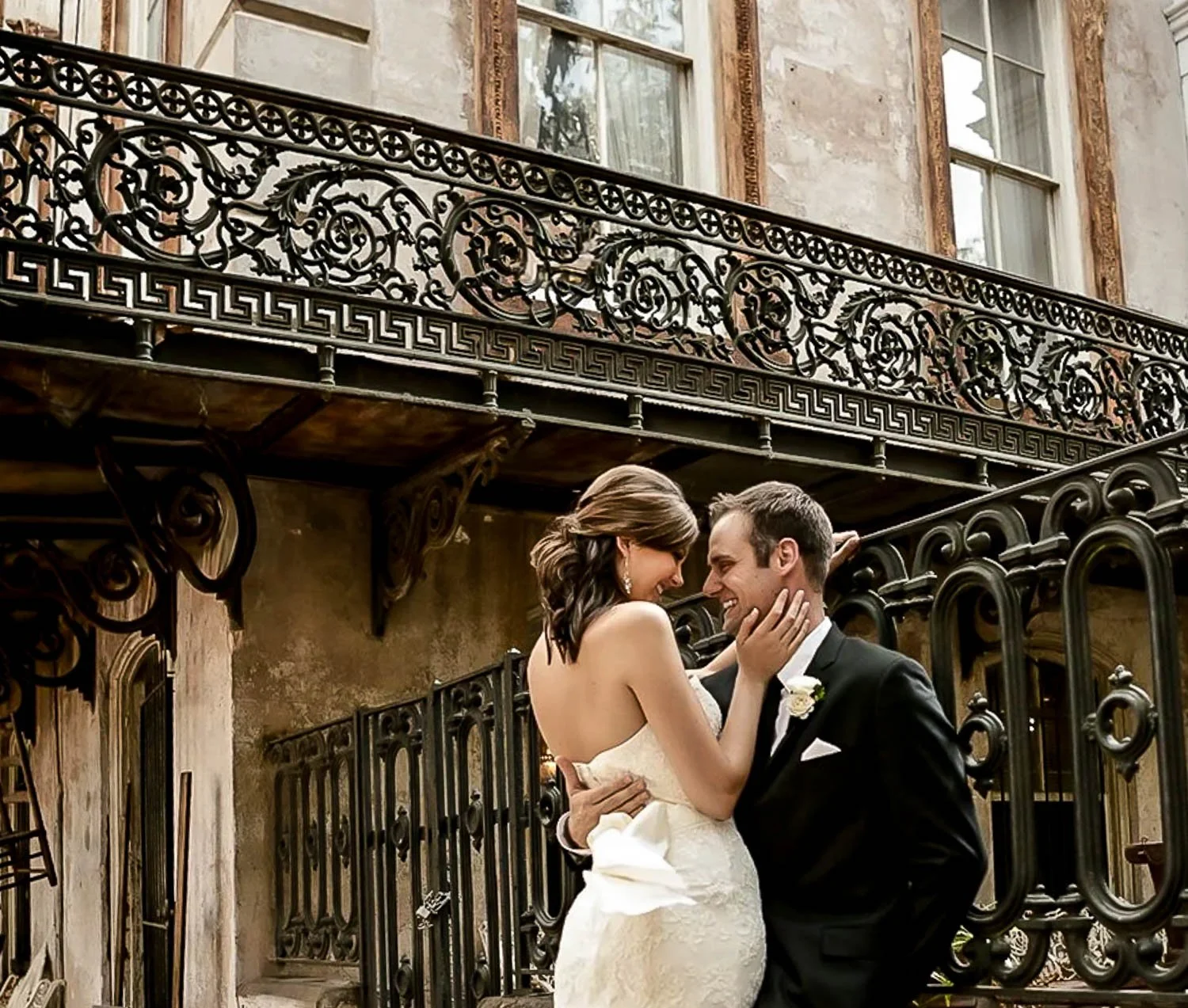 A bride and groom sharing an intimate moment, smiling and holding each other, outside near ornate black wrought iron gates and a building with large windows.