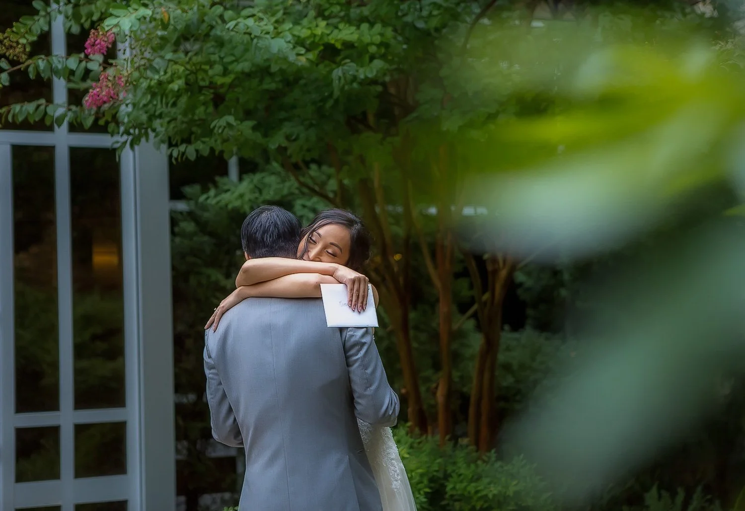 A couple hugging outdoors in a garden, with the woman holding a white envelope, surrounded by greenery and pink flowers.