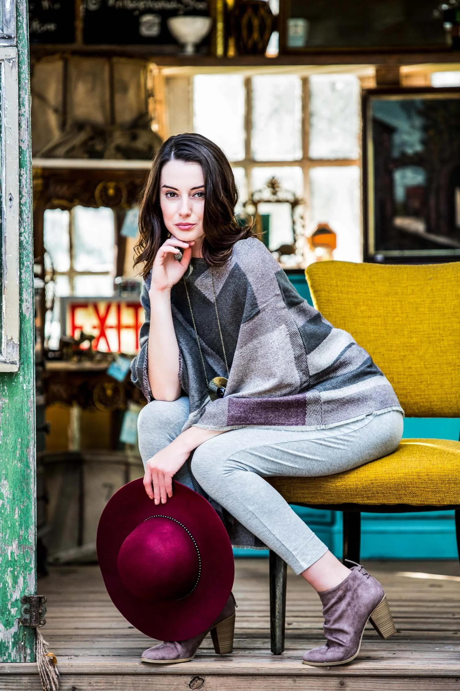 A woman with dark hair and fair skin is sitting on a yellow chair in a rustic, vintage room. She is wearing a gray plaid poncho, light gray pants, and taupe ankle boots, and is holding a burgundy wide-brim hat and resting her chin on her hand while l