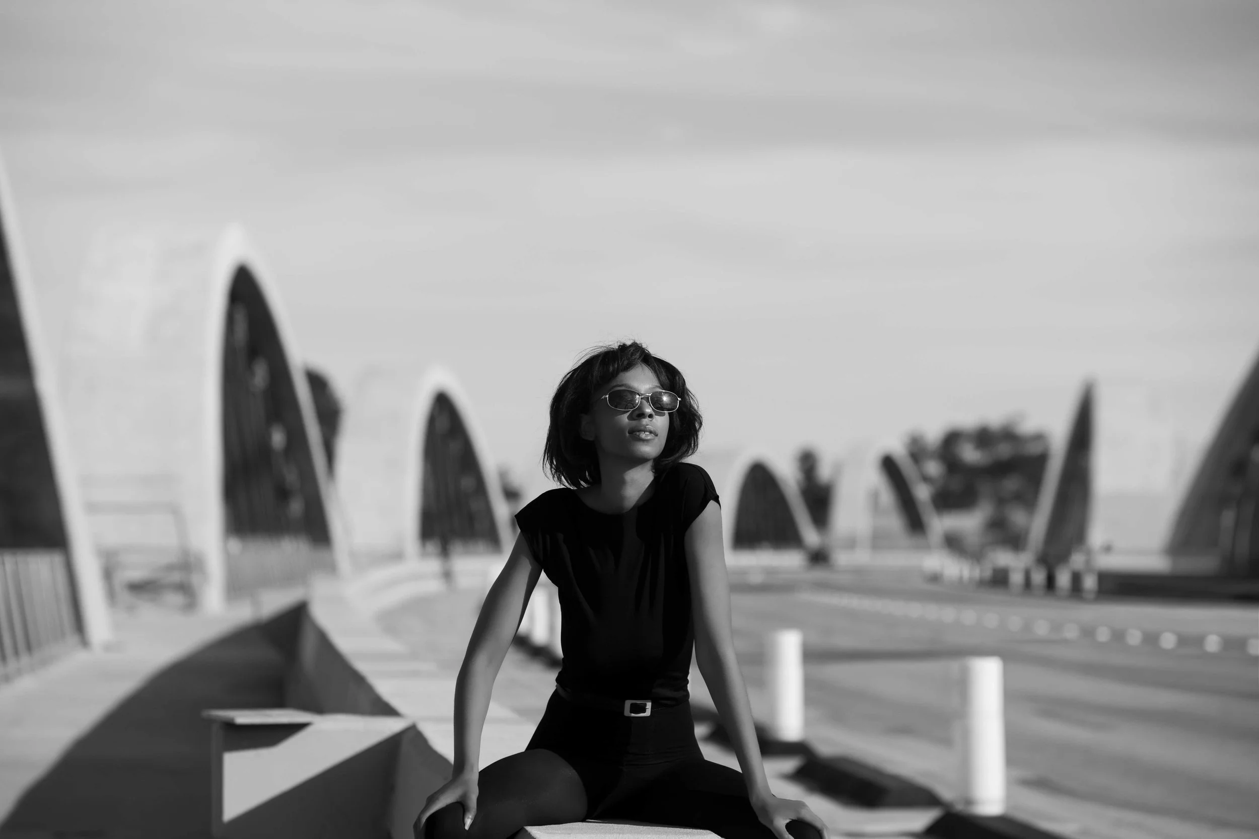 A woman wearing sunglasses and a black dress sits on the ground at a beachside promenade, with large white arches and empty chairs in the background, under a cloudy sky.