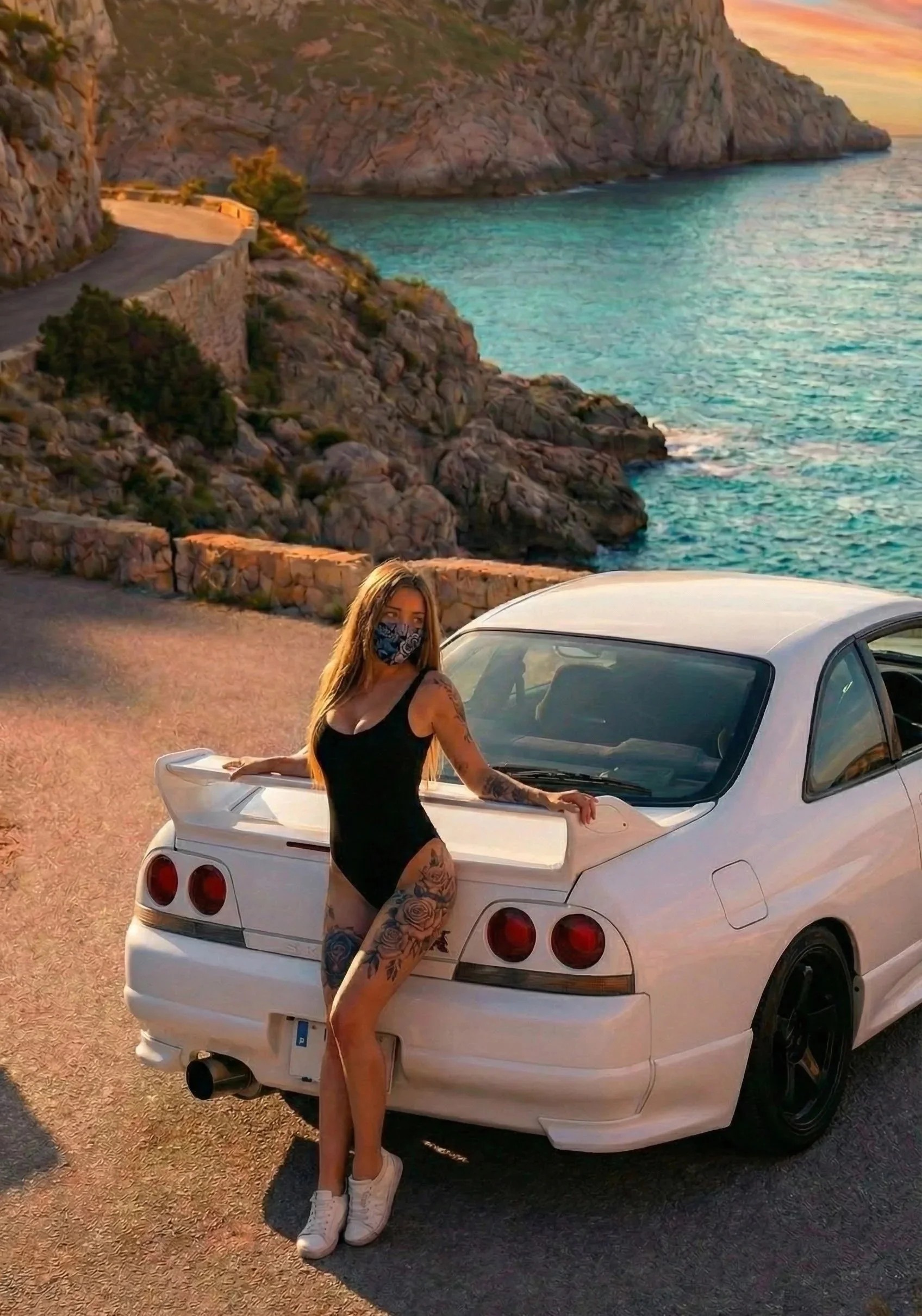 A woman with tattoos wearing a black swimsuit and a face mask stands next to a white sports car by the coast during sunset, with rocky cliffs and the ocean in the background.