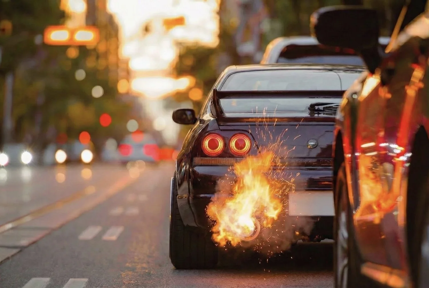 A black sports car with a large rear spoiler is on fire at the back, with flames and sparks coming from the exhaust, on a city street during dusk with blurred traffic and lights in the background.