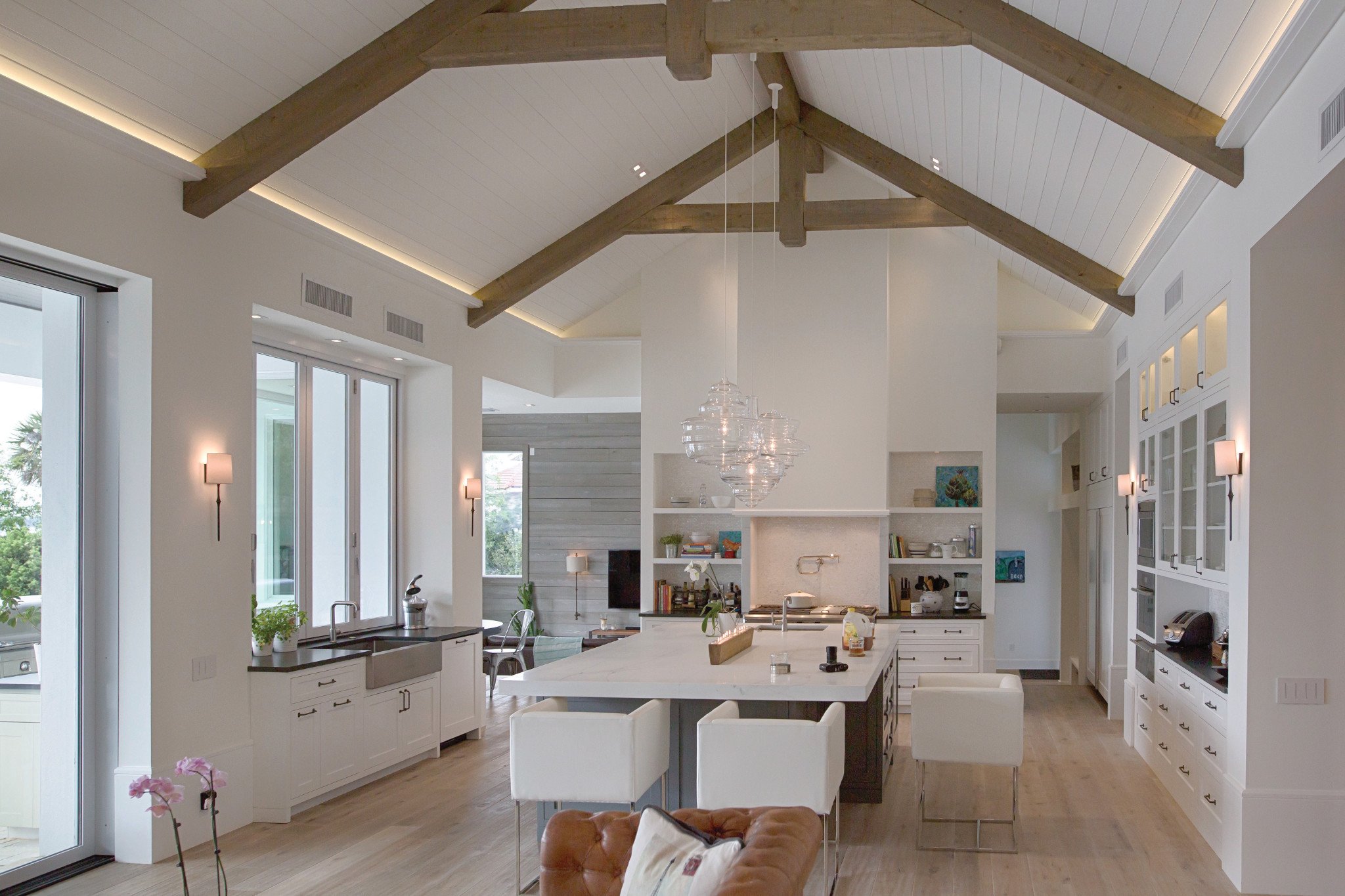Open-concept kitchen with high vaulted ceilings, wooden beams, white walls, and a large island with seating. Features include wall-mounted lights, large windows, and built-in cabinetry, with a view of a cozy living area and Clara Shades. 