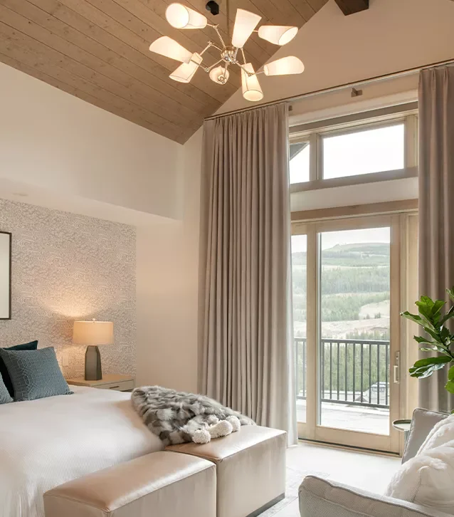 Cozy bedroom with a large bed, gray and white pillows, a gray fur throw, a beige ottoman at the foot, beside a nightstand with a lamp, large beige automated drapery curtains, a sliding glass door leading to a balcony, and a view of mountains outside.