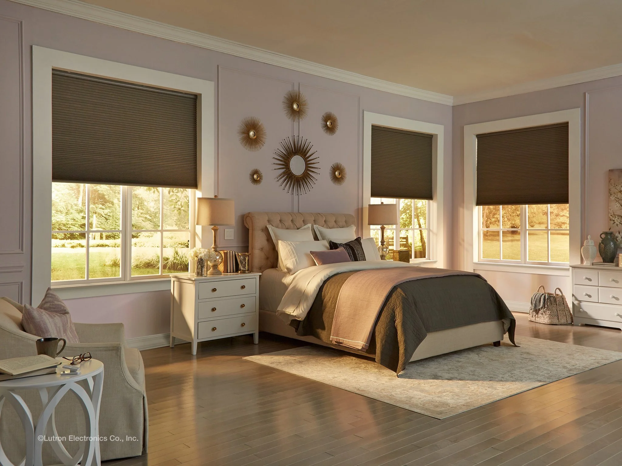 A bedroom with a beige bed, nightstands, lamps, and windows with dark blinds. The room has light-colored walls, wooden floors, and minimal decorative items.