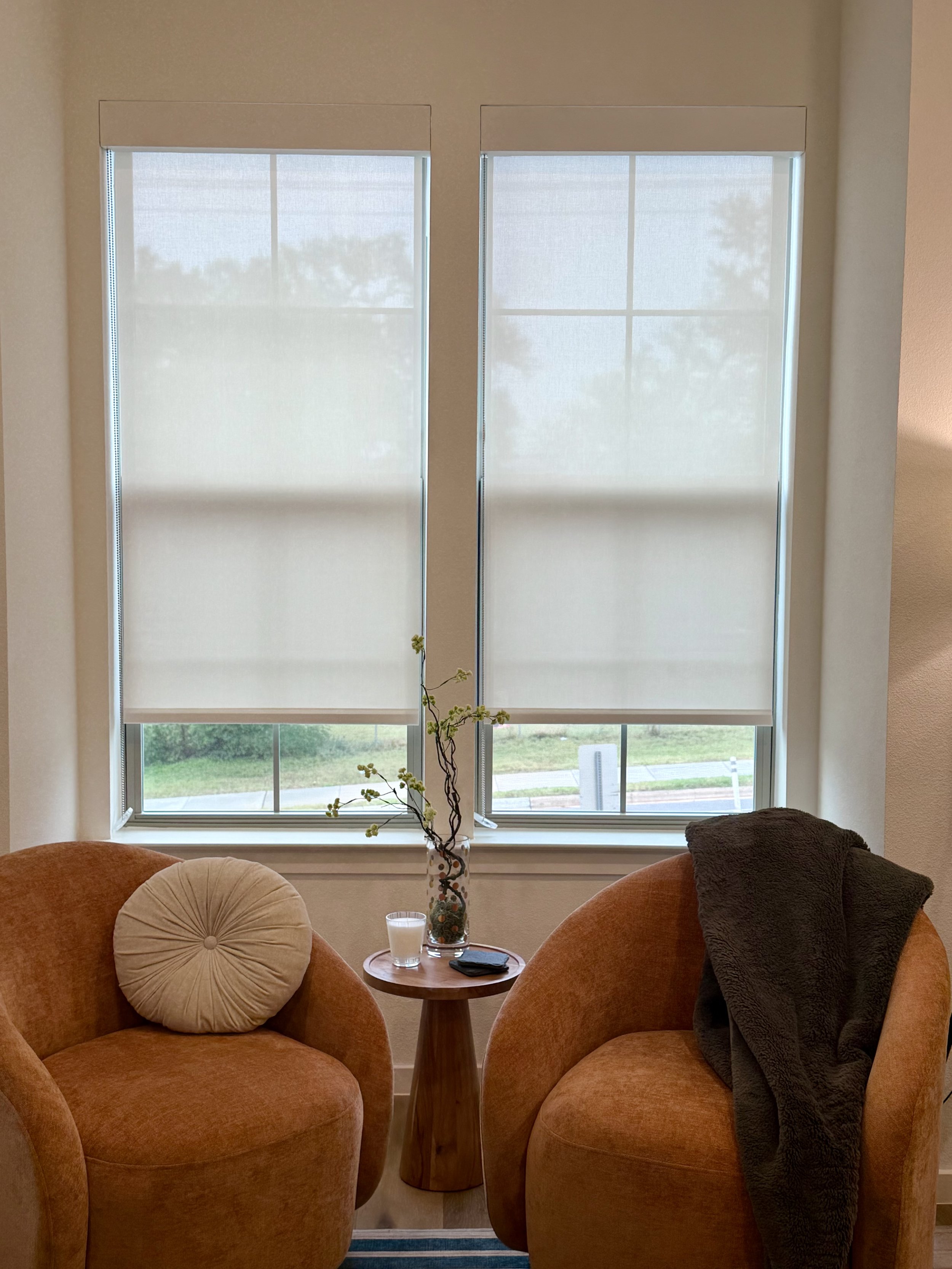 Two cream roller shades professionally installed on side-by-side living room windows in East Austin, with warm amber armchairs and a small round wood accent table.
