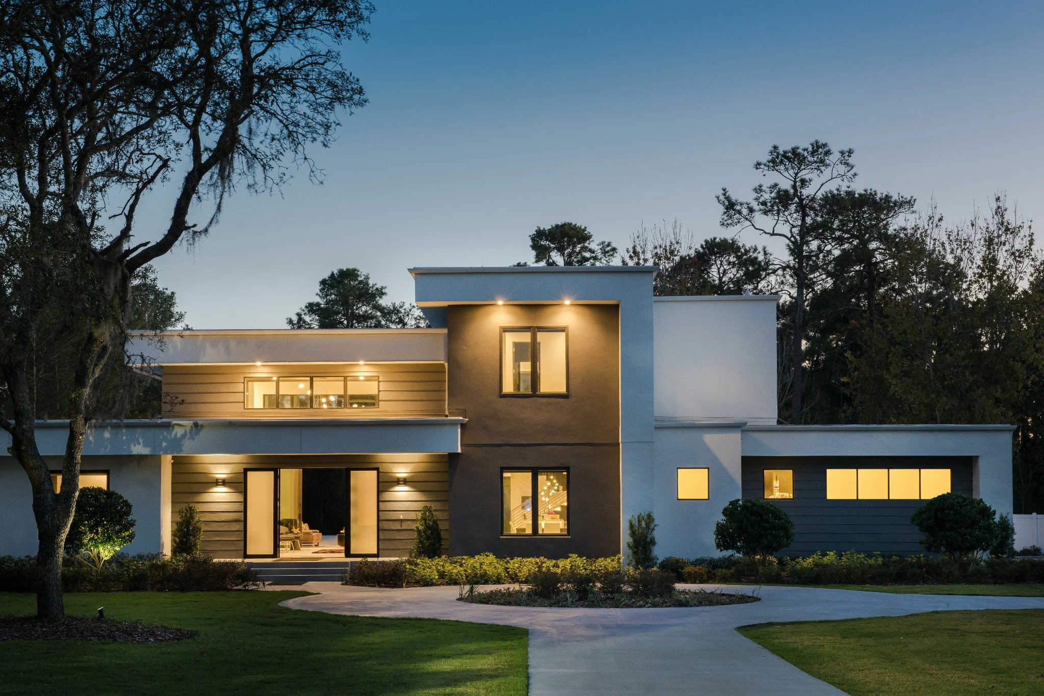 Modern two-story house with large windows, surrounded by a well-manicured lawn, trees, and bushes, illuminated at dusk with automated smart lighting.
