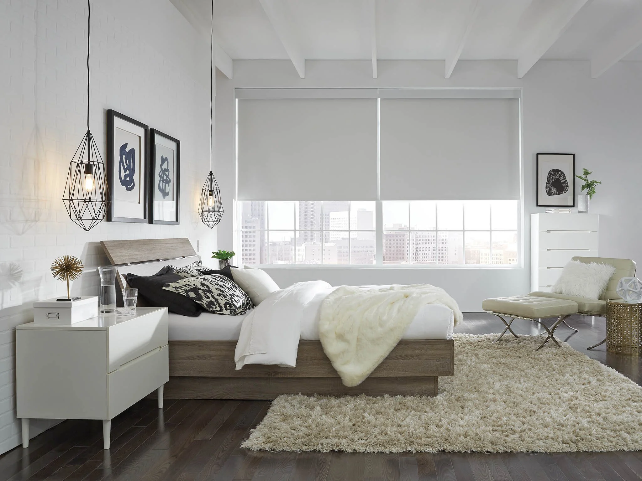 Modern bedroom with white walls, large windows with blinds, wooden bed with black and white bedding, beige rug, white nightstand with decor, lounge chair, and minimalistic decor.