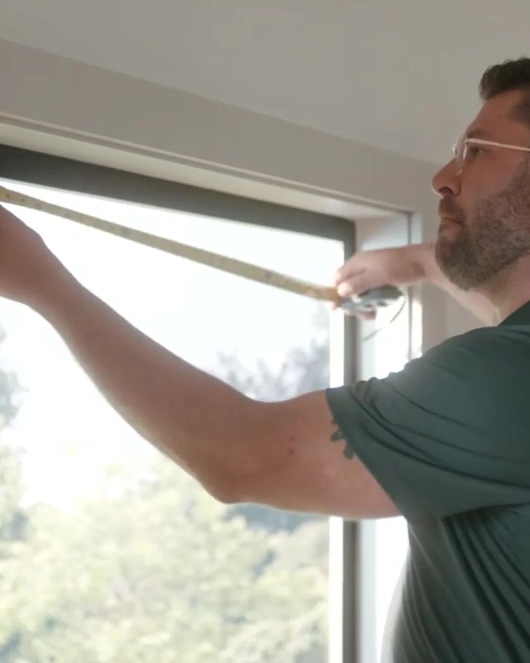 A man with glasses, a beard, and tattoos on his right arm is cleaning a window with a squeegee.