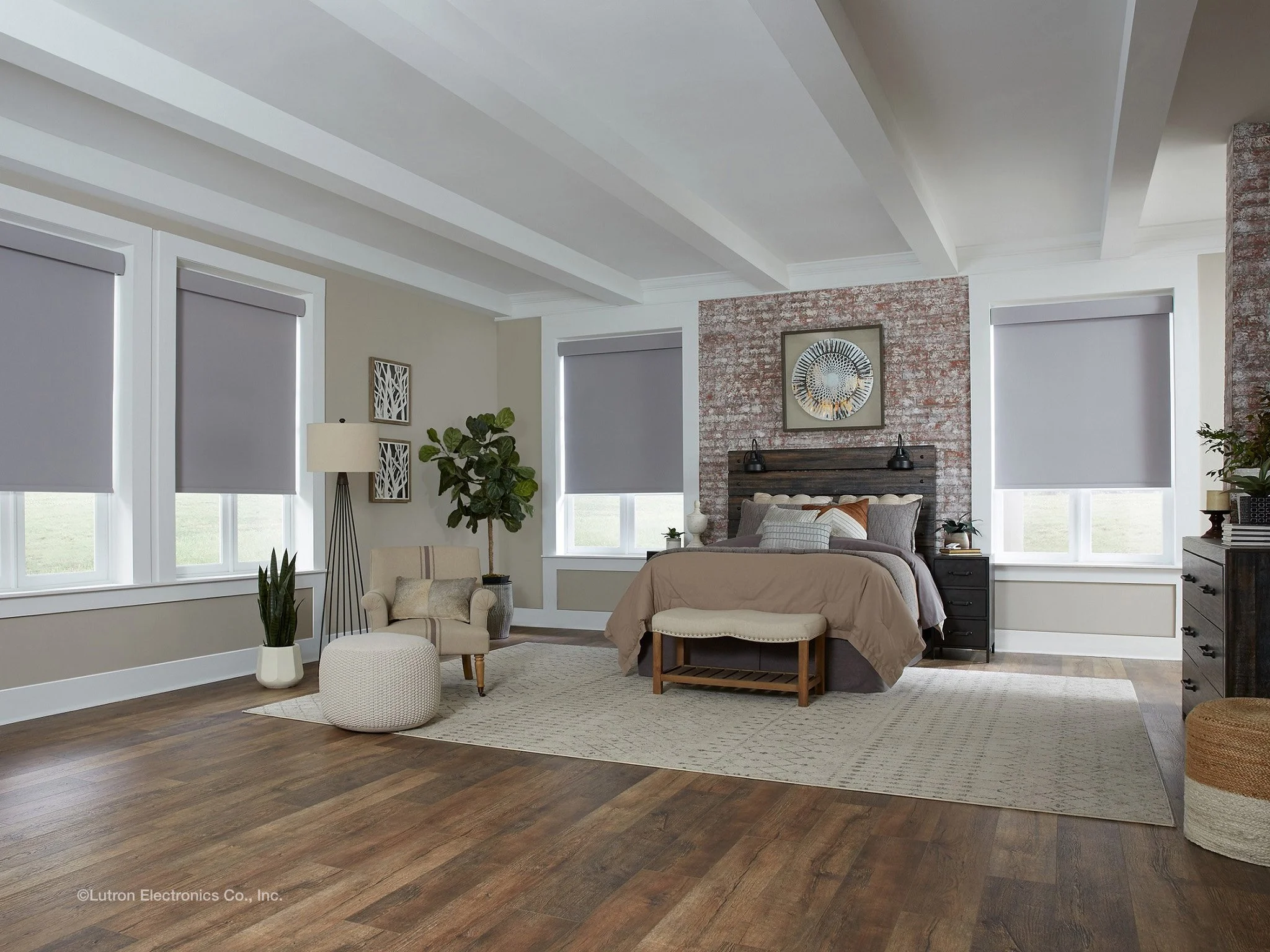 A spacious bedroom with a bed, nightstands, and framed artwork on an exposed brick wall. There are three windows with gray motorized Lutron window shades, a beige armchair with a floor lamp, and a large area rug on wooden floors.