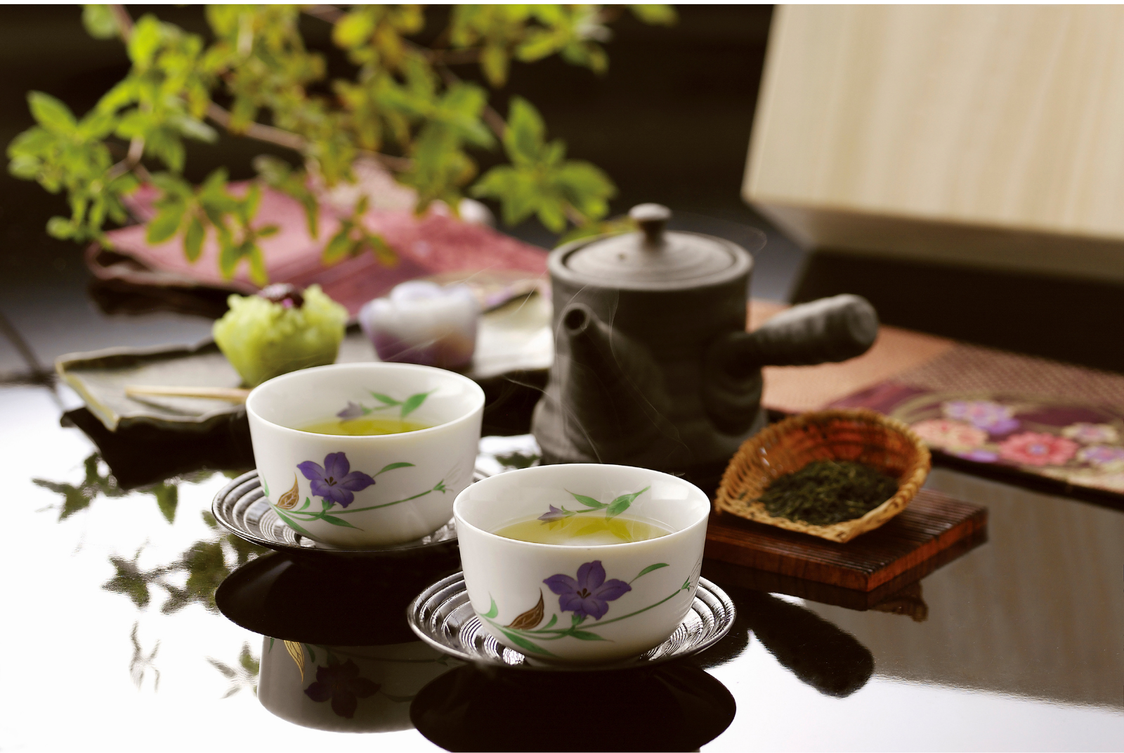 Two white cups with purple flower designs filled with hot tea, placed on a black reflective table. Behind the cups, there are a black teapot with steam, a tray with dried tea leaves, and various decorative items including a green fruit, a purple object, and a plant with green leaves in the background.