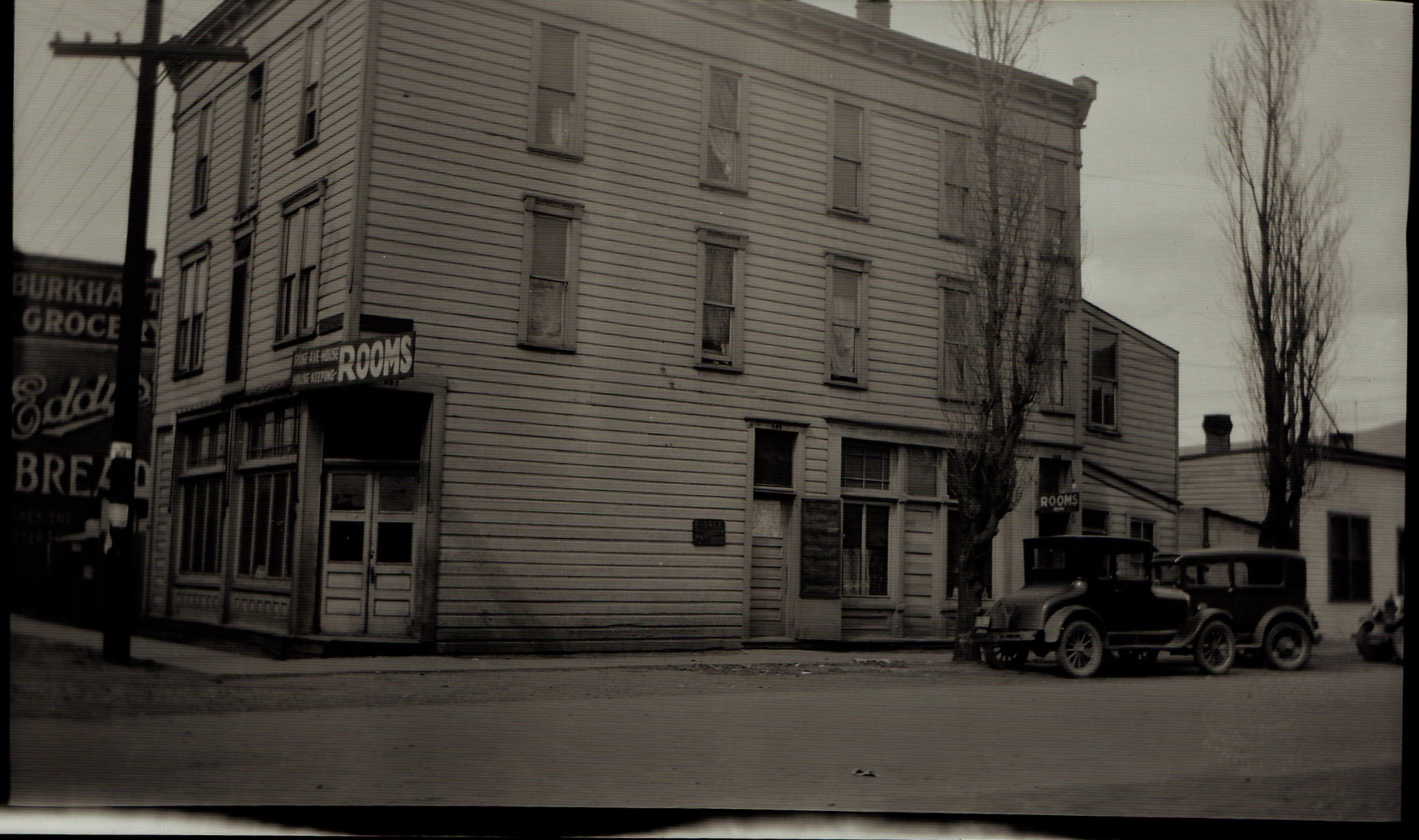 Black and white photo of a three-story brick building with signs indicating it is a rooming house and bakery. Trees are visible, and an old automobile is parked in front.