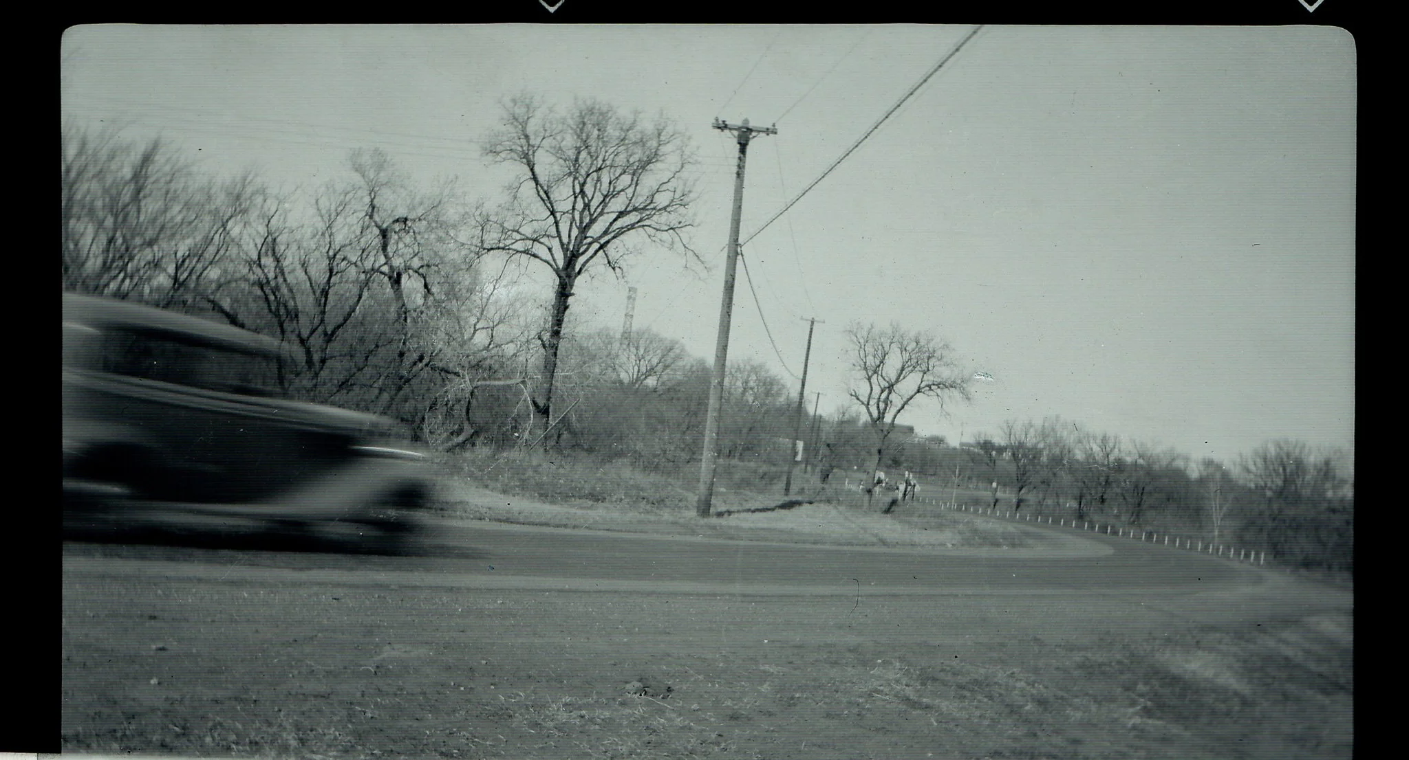 Black and white photograph of a road with a moving car on the left side, power lines, leafless trees along the roadside, and a cloudy sky.