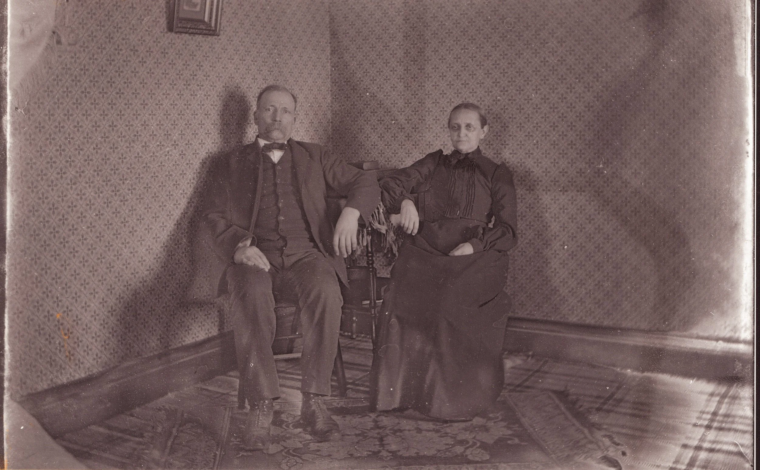 An old black and white photograph of a man and woman sitting together in a room with patterned wallpaper. The man is dressed in a suit with a bow tie, and the woman is wearing a long dark dress. They are sitting on chairs, with the woman's arm restin