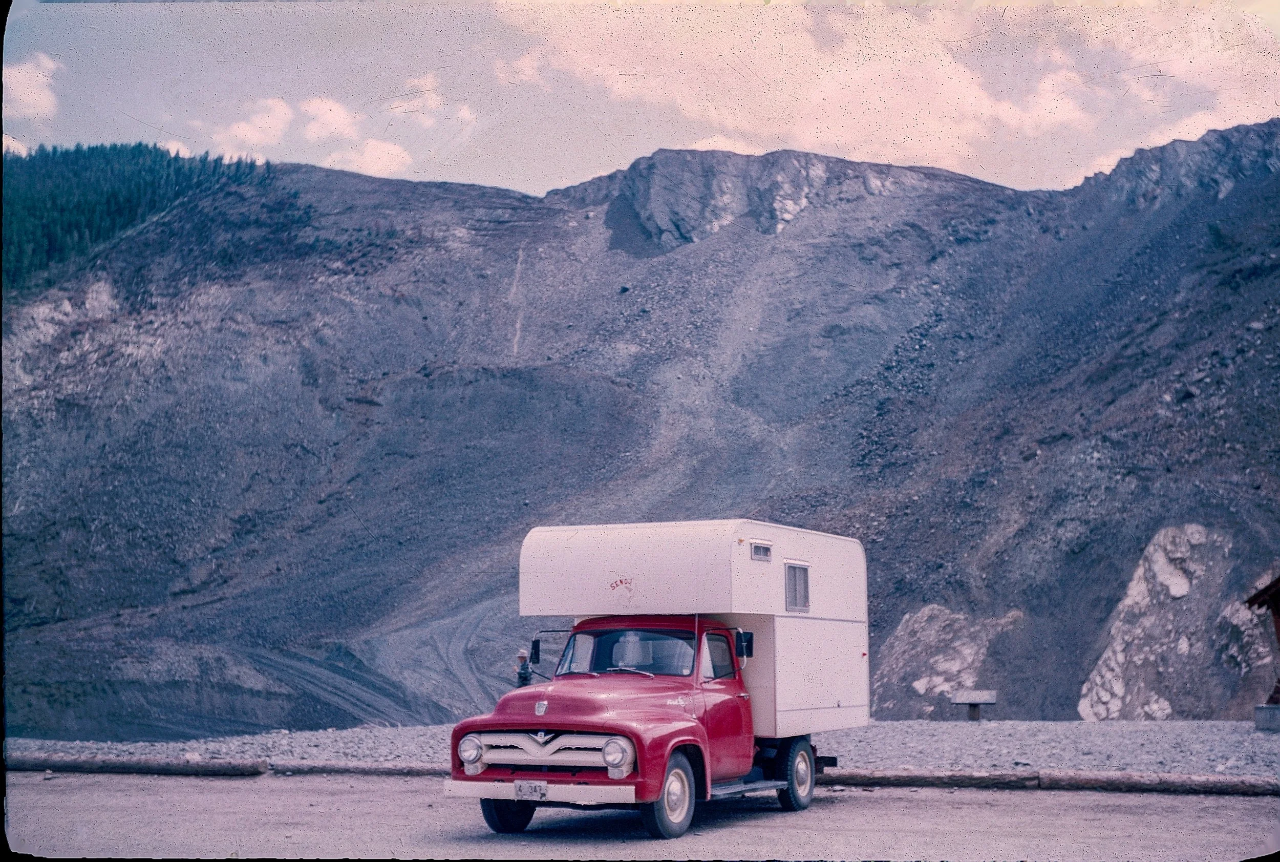 A red truck with a white camper attached parked outdoors on a dirt surface against a backdrop of a rocky mountain landscape under a sunrise or sunset sky.